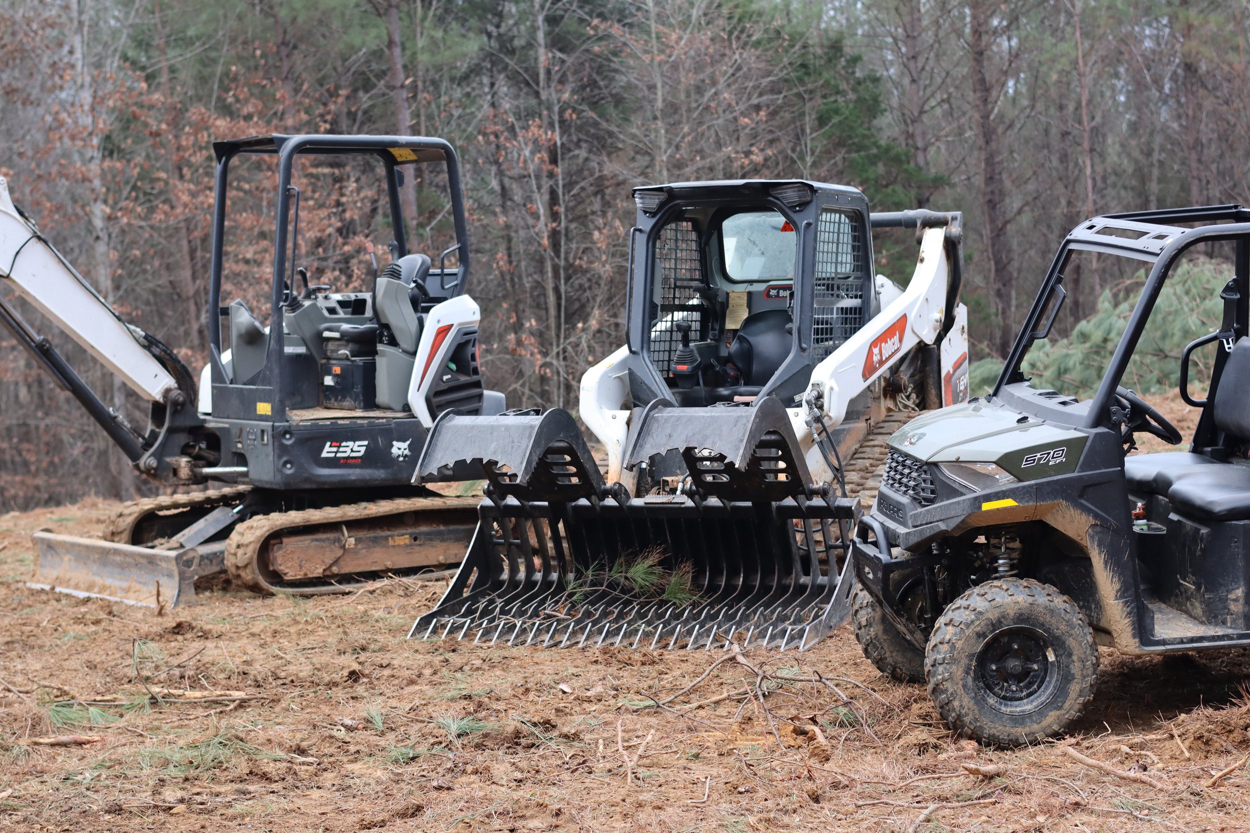 Three small construction vehicles on dirt ground in a wooded area