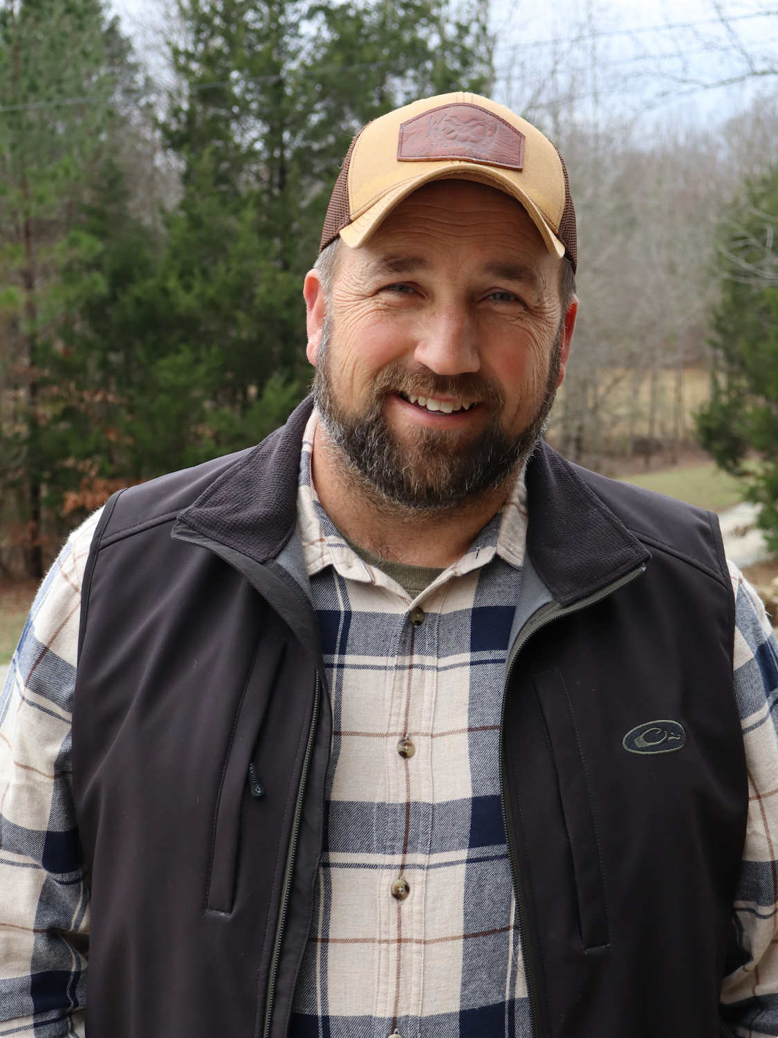 A smiling man outdoors during daytime, wearing a tan cap, plaid shirt, and black vest, with a background of trees and a hint of cloudy sky.
