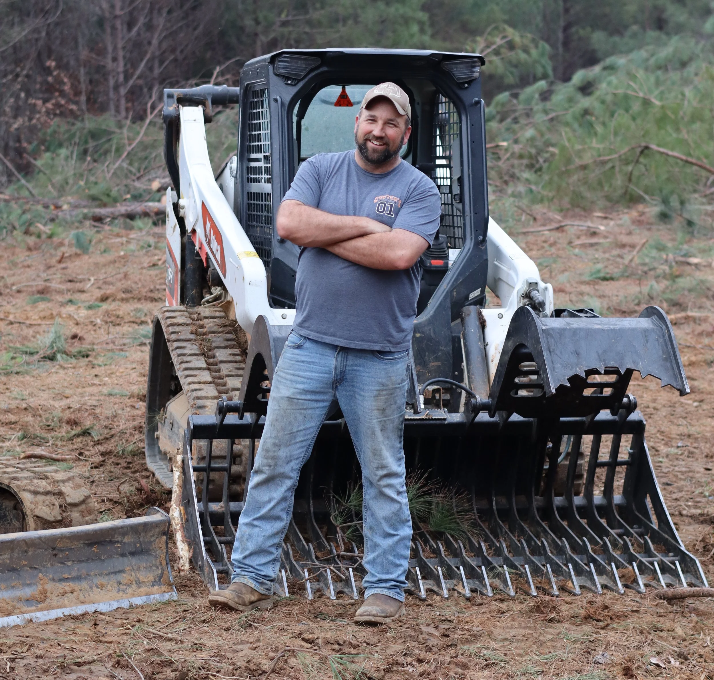 A man with a beard and wearing a baseball cap, t-shirt, and jeans standing with arms crossed in front of a skid-steer loader on a dirt field surrounded by trees.