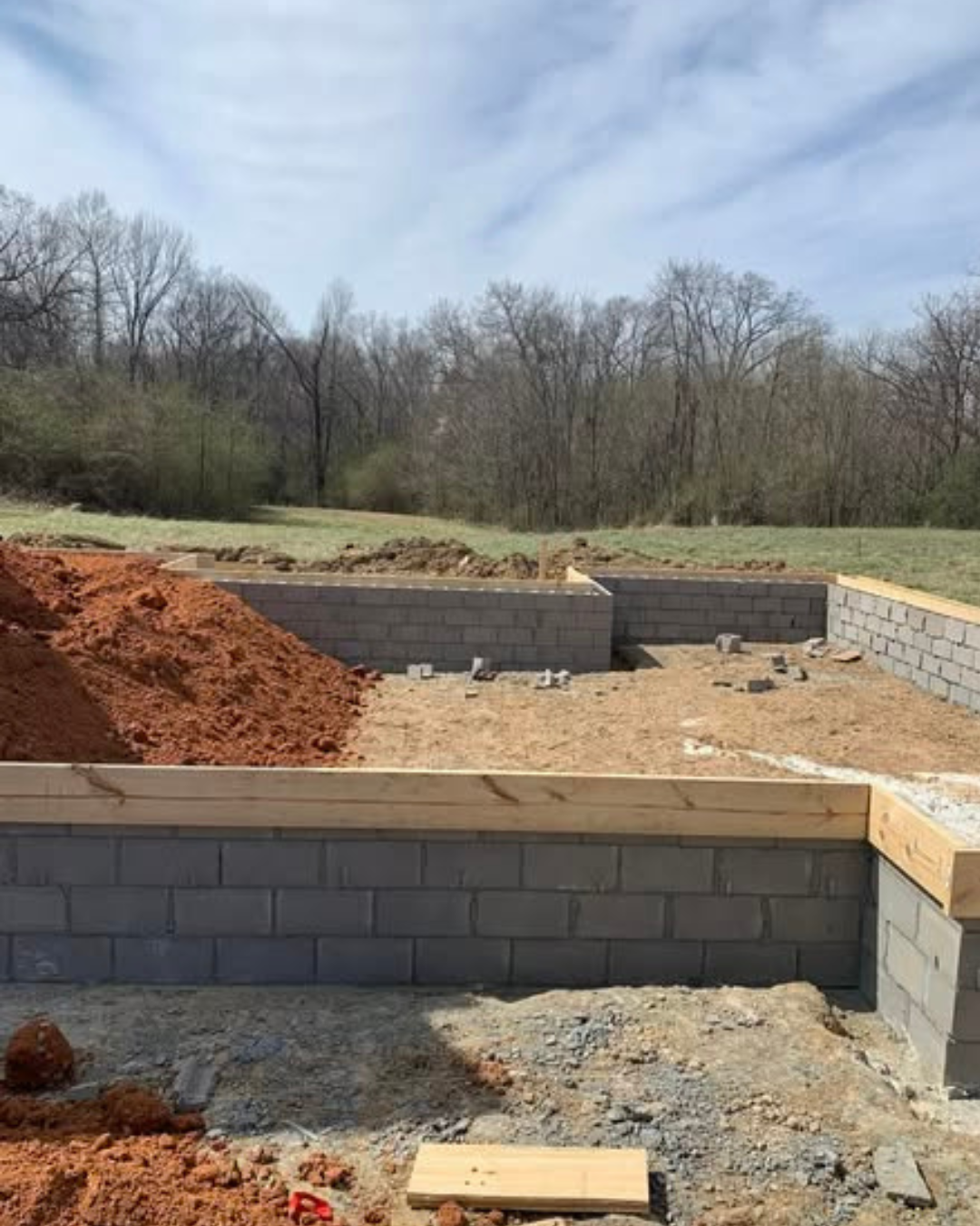 Construction site with brick walls being built outdoors on a grassy field with trees and a cloudy sky in the background.