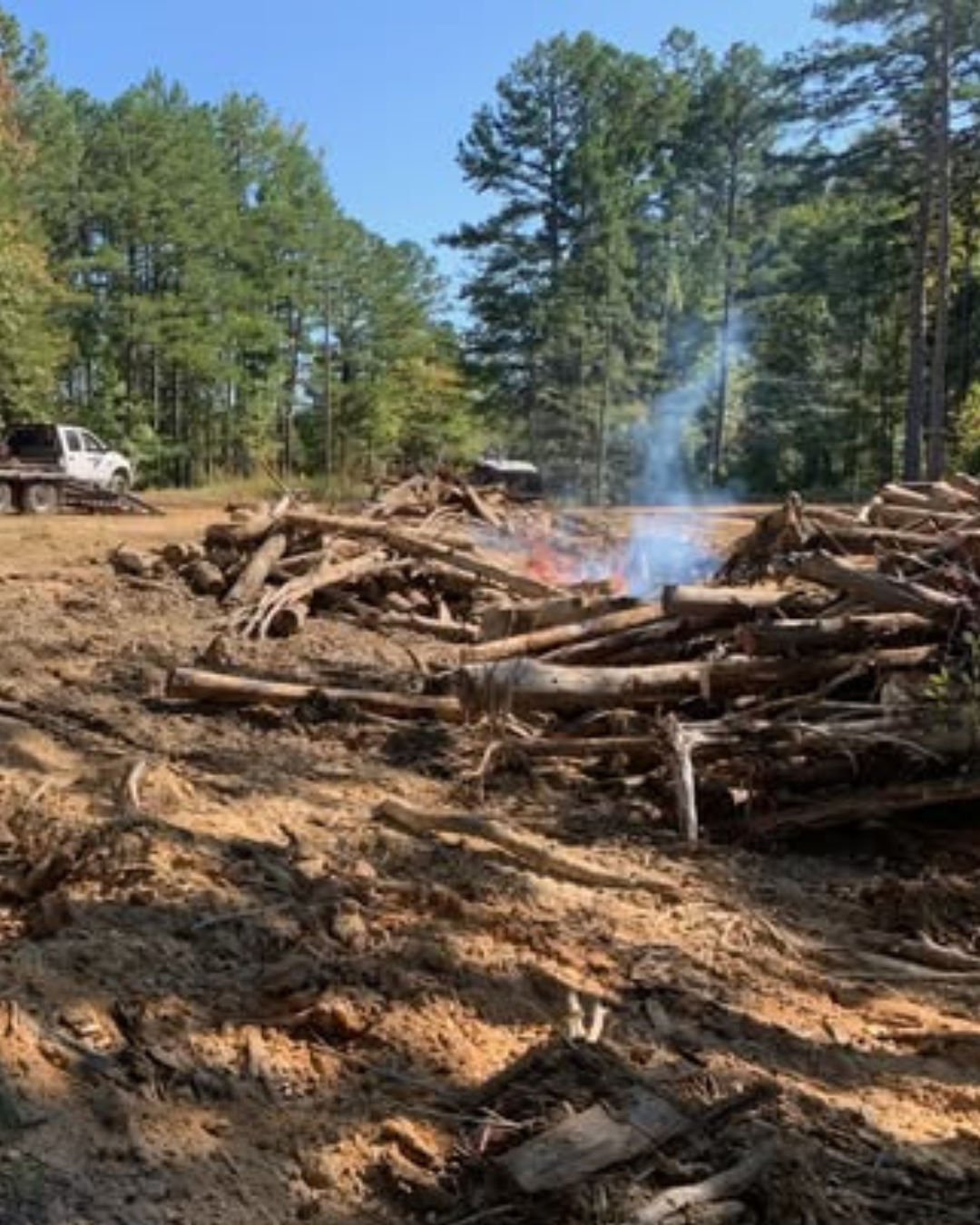 A pile of wooden logs and splinters on dirt ground in a forest clearing with some smoke and small flames, and a white truck parked in the background among tall pine trees.