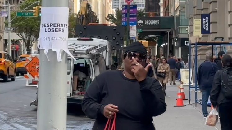 A woman wearing sunglasses and a black cap, standing on a city sidewalk, holds a red bag and smokes a cigarette. Behind her, there is a damaged vehicle, construction cones, and pedestrians.