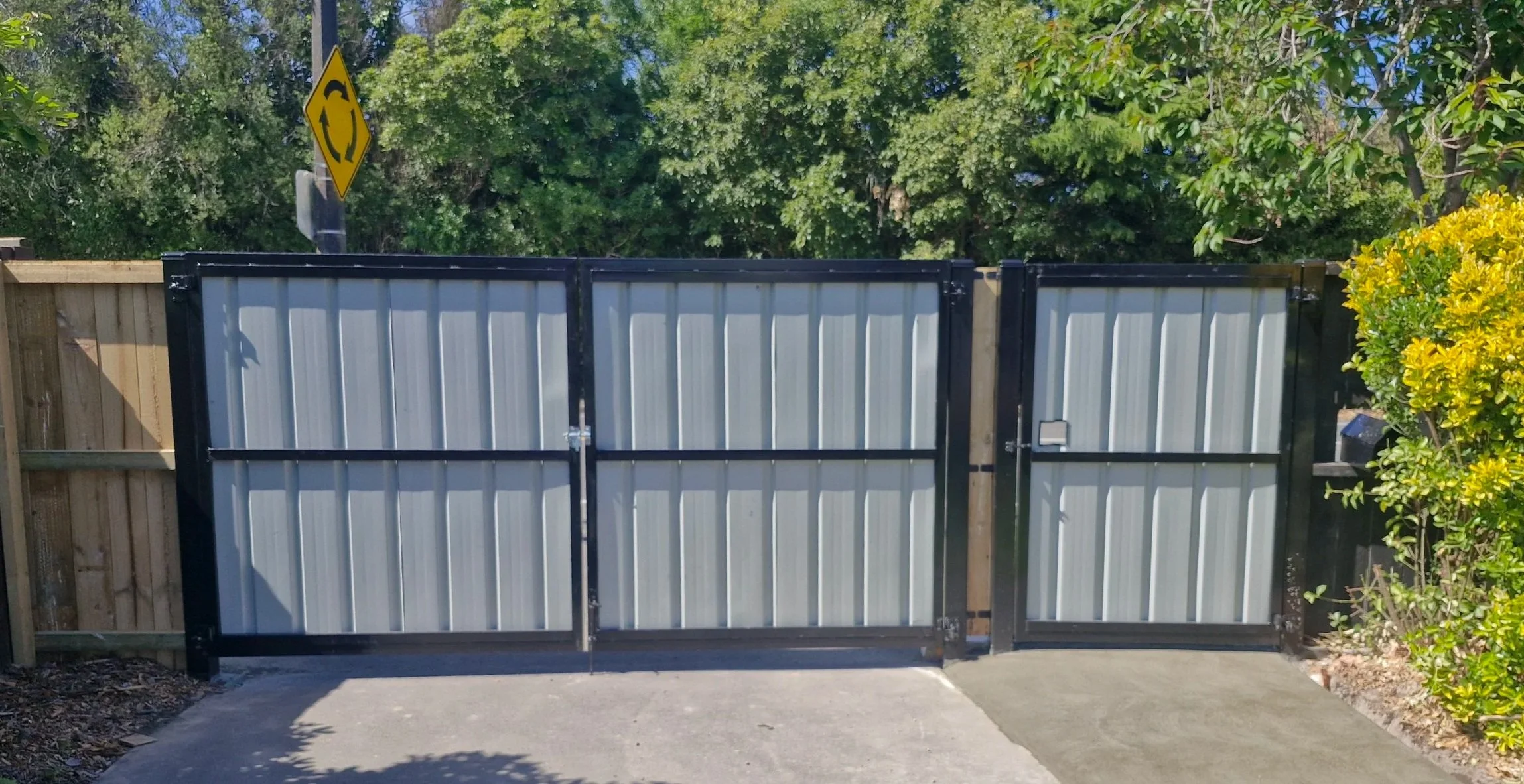 A metal sliding gate with a black frame, set in a wooden fence with trees and bushes in the background.
