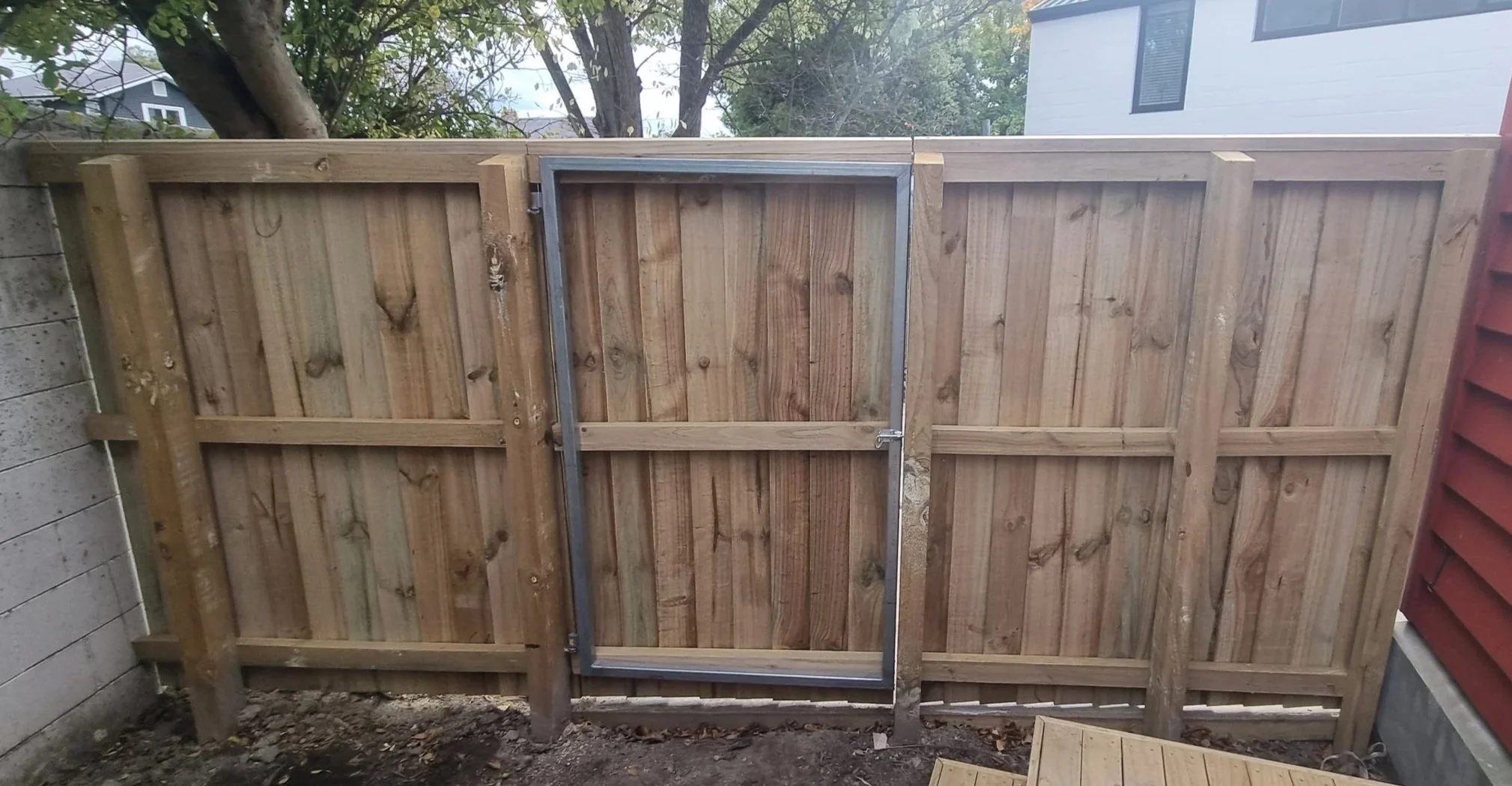 A wooden fence with a metal frame gate installed in a backyard, with trees and a house visible in the background.