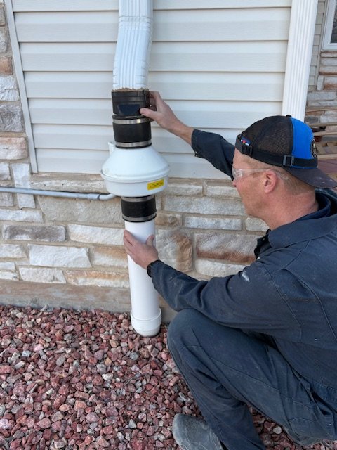 A man wearing a black cap and jacket adjusts a drainage pipe on the side of a house with white siding and a stone foundation, using his hands.