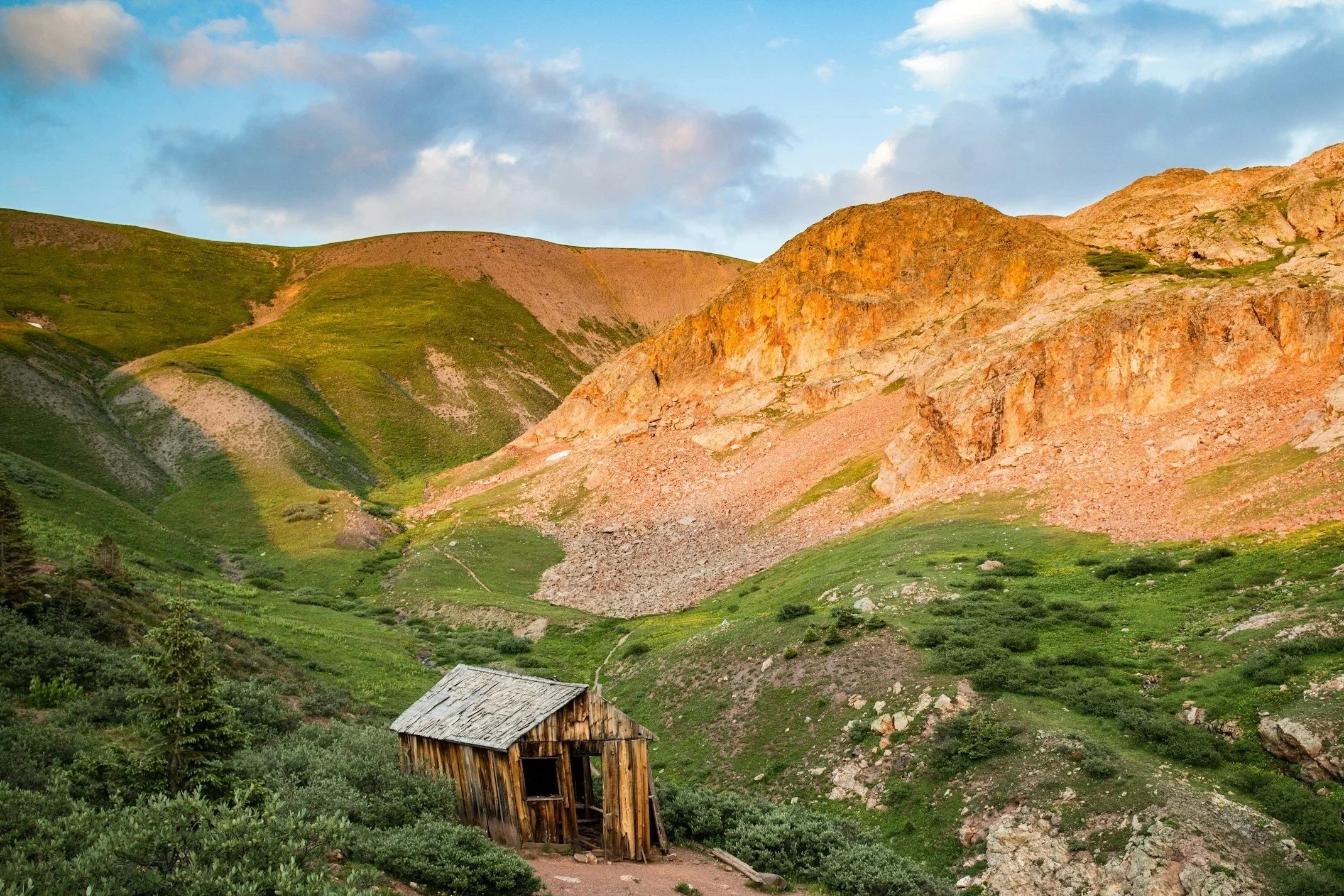 A small, weathered wooden cabin is situated in a lush green valley with rolling hills and colorful rocky mountains in the background under a partly cloudy sky.