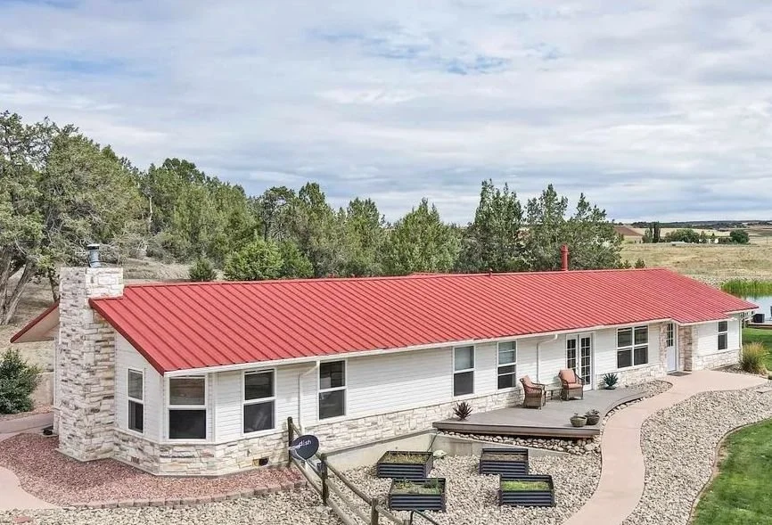 Single-story house with white siding, stone accents, a red metal roof, and a stone chimney, surrounded by a landscaped yard with pathways and outdoor seating.