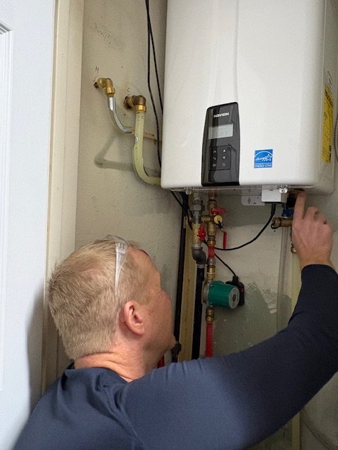 A man working on a wall-mounted white water heater or boiler, reaching towards it with tools in his hand.