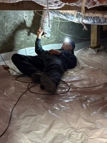 A man lying on the floor underneath a construction ceiling, holding a flashlight in a basement or crawl space with insulation and exposed piping.
