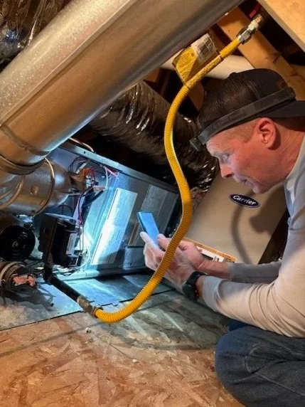 A technician inspecting an HVAC furnace under the house with a flashlight and a smartphone.