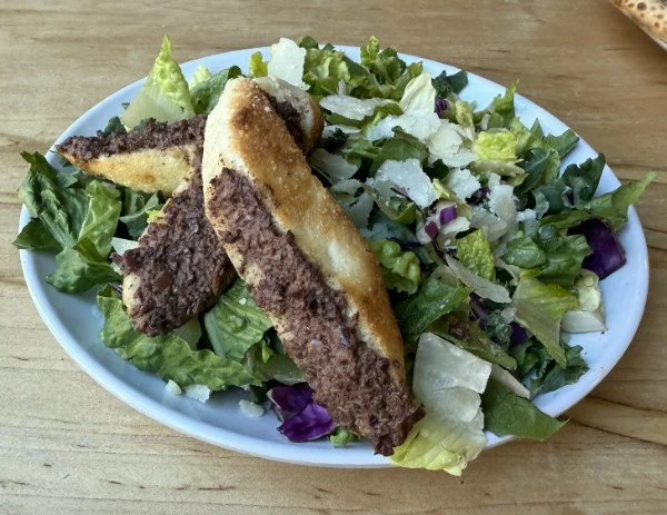 A white oval dish containing a salad with mixed greens and chopped vegetables, topped with two slices of breaded and fried meat, served on a wooden table.