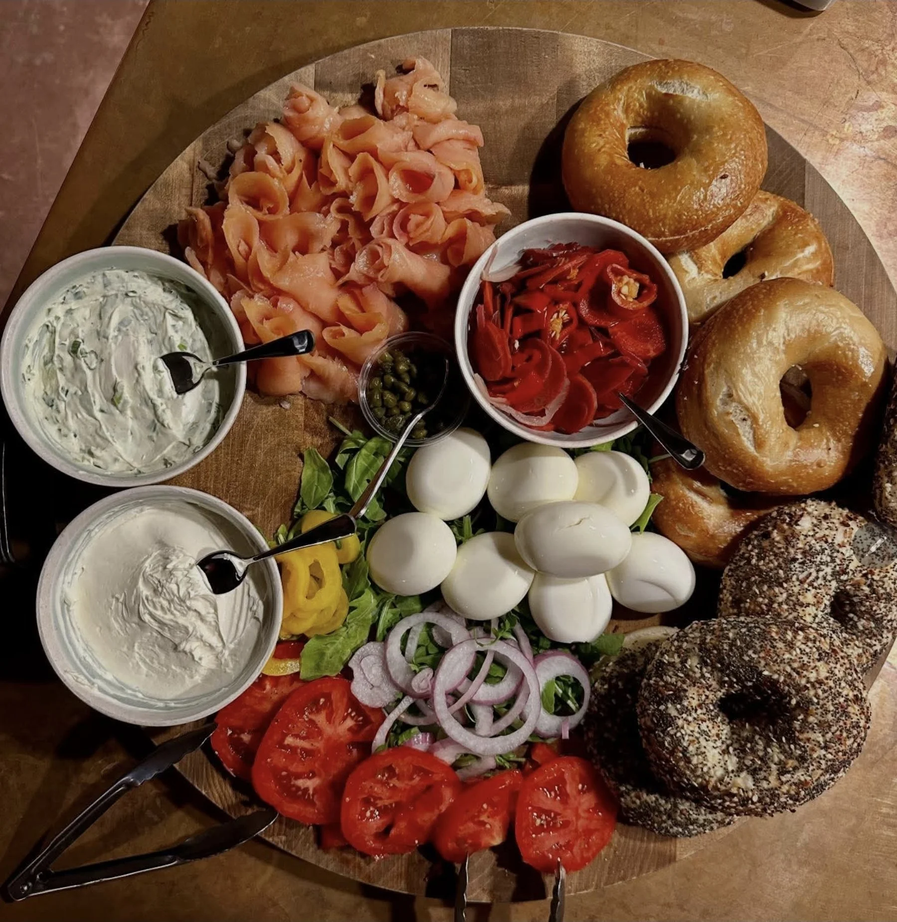 A wooden round serving board with various breakfast items including bagels, sliced tomatoes, sliced onions, fresh mozzarella balls, smoked salmon, pickles, and bowls of cream cheeses and smoked red peppers, with some greens and sliced yellow peppers.