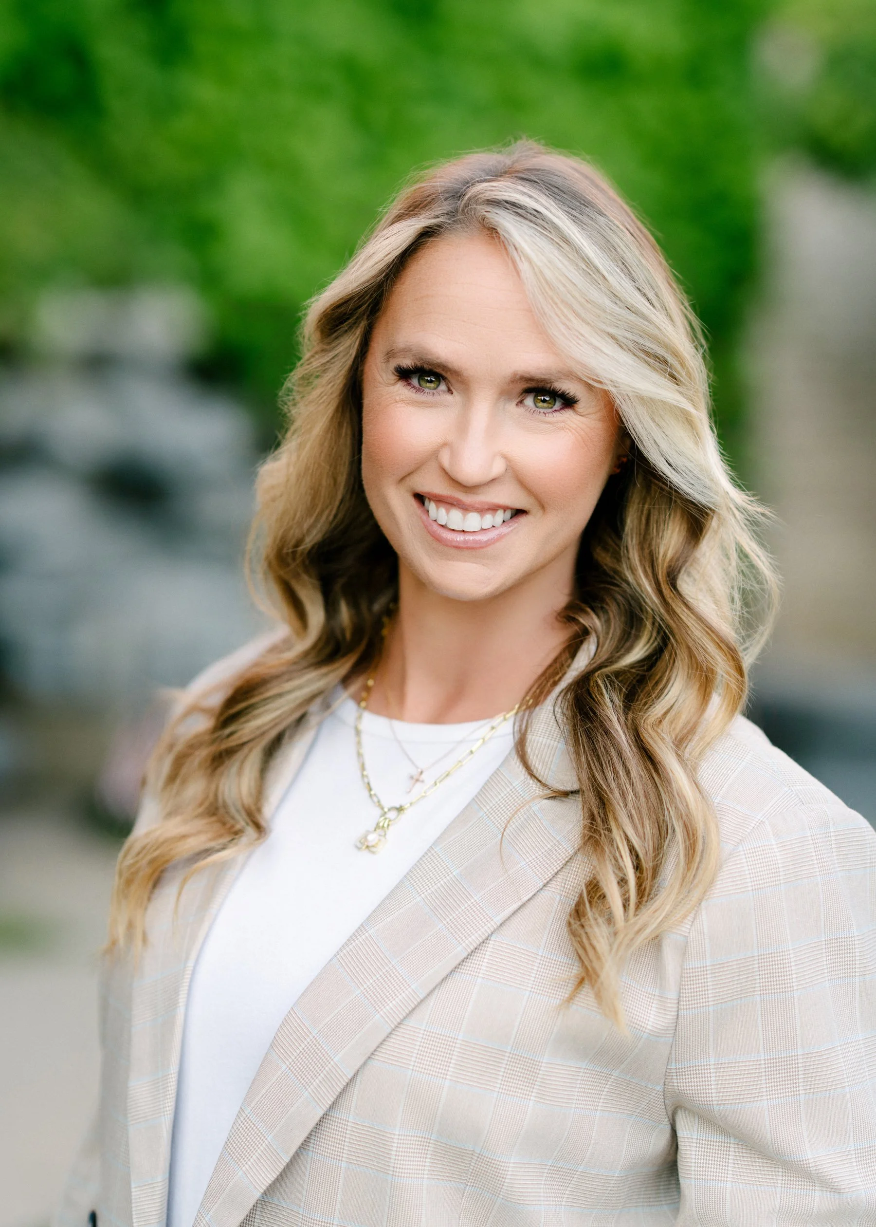 A woman with blonde, wavy hair and green eyes smiling, wearing a light beige checkered blazer, white top, and layered gold necklaces, standing outdoors with blurred green trees in the background.