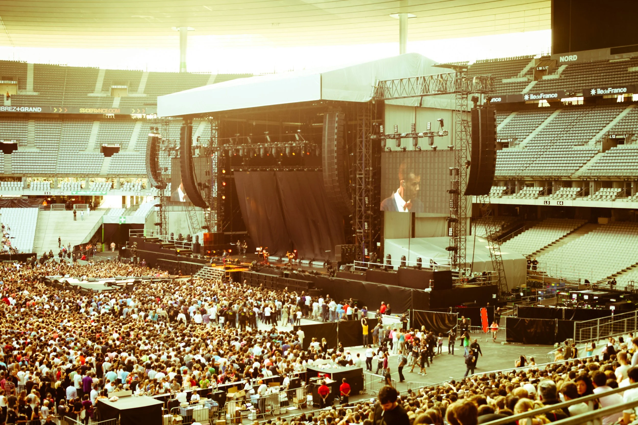 Large indoor stadium filled with a crowd of people attending a concert or event. Stage setup with big screens and lighting equipment, some audience members are standing and others seated, with some walking around.