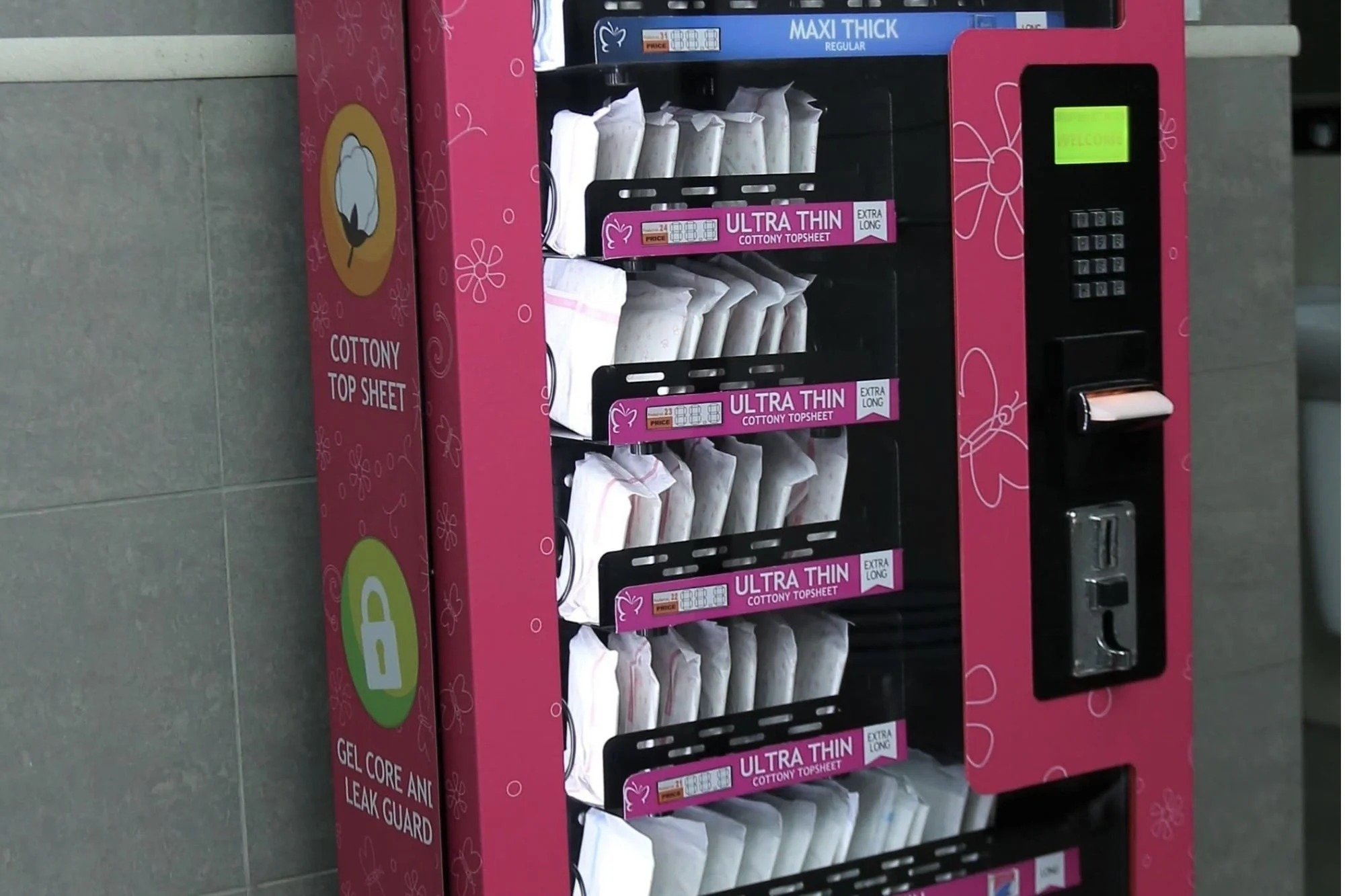 Pink vending machine with cotton topsheets and leak guard pads, labeled as ultra thin, extra long, cottony top sheet, with a keypad and card payment slot.
