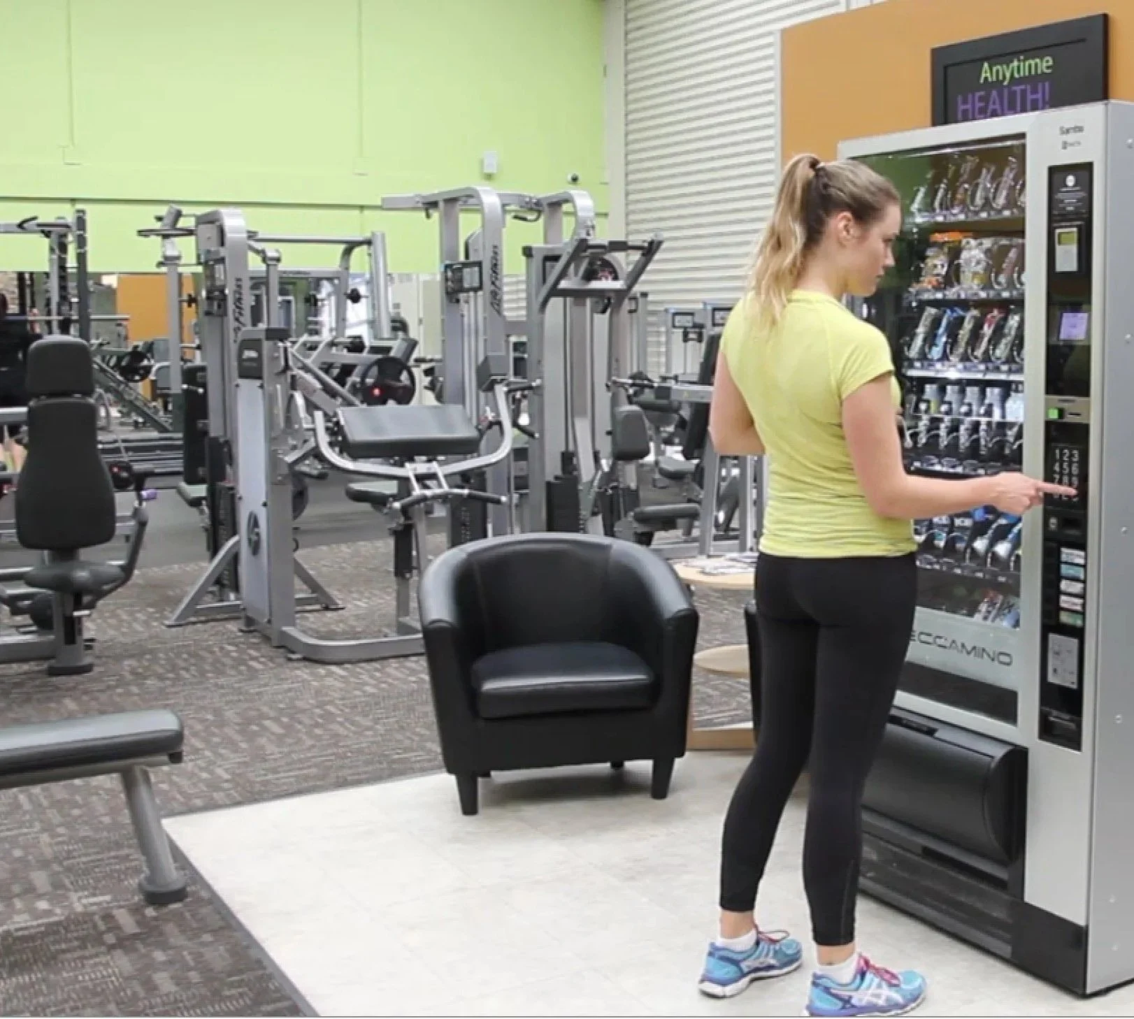 A woman in a yellow shirt and black leggings uses a vending machine in a gym. The gym has various exercise machines and a black chair nearby.