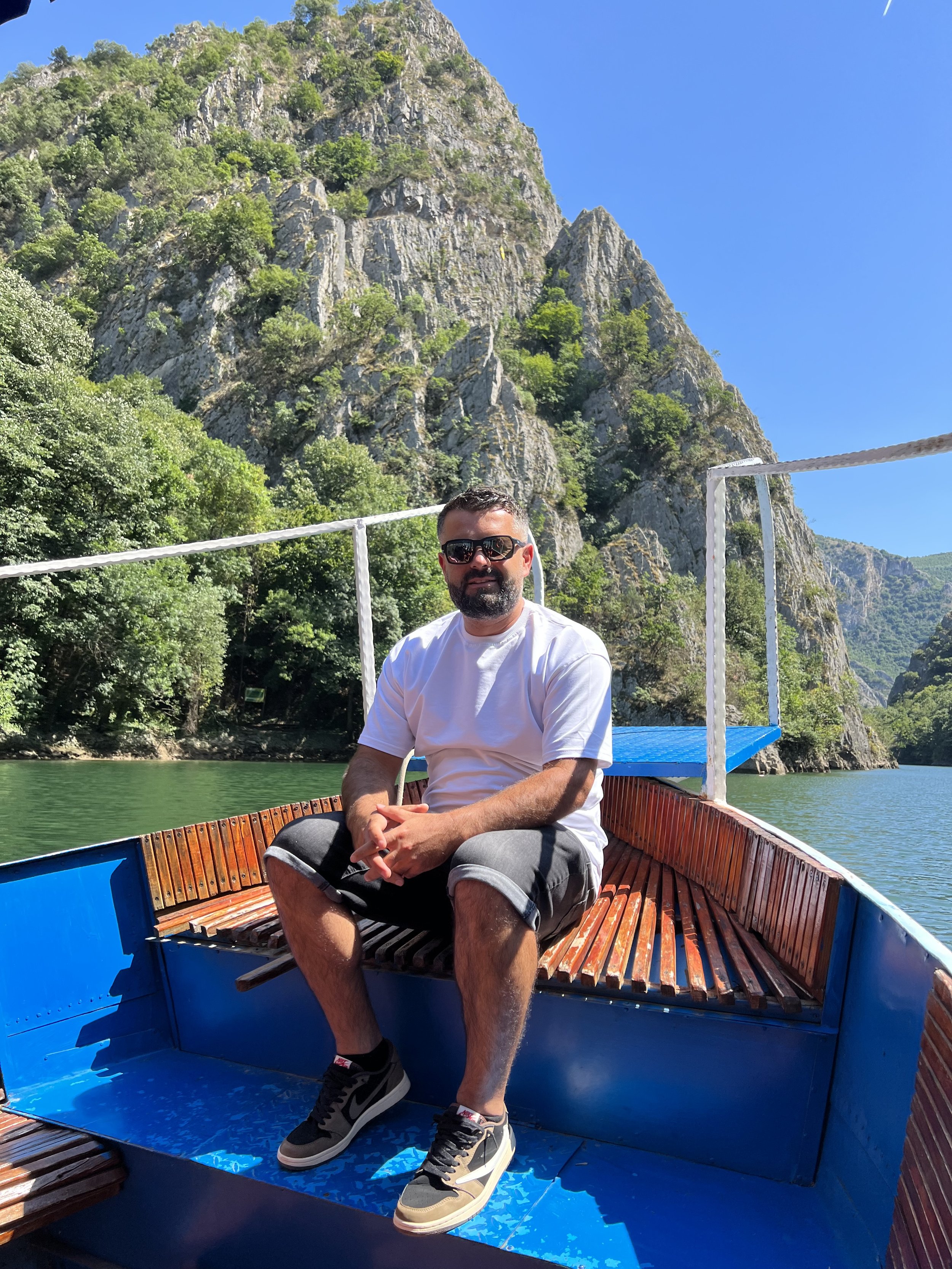 A man with sunglasses, a white t-shirt, shorts, and sneakers sitting on a boat with a scenic mountain and greenery in the background.