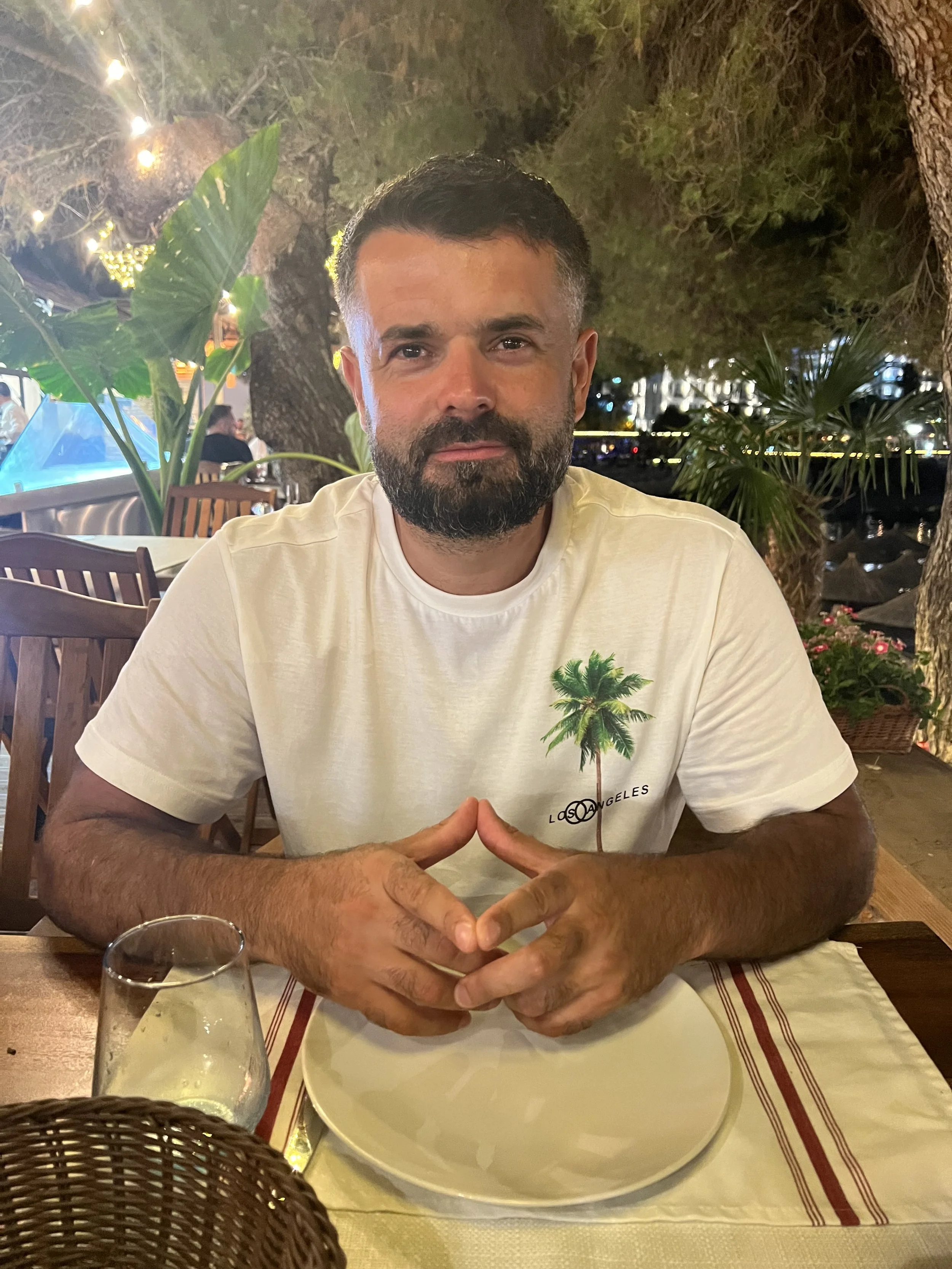 A man with dark hair and a beard sitting at a table outdoors at night, wearing a white T-shirt with a palm tree graphic and the text 'Los Angeles'.