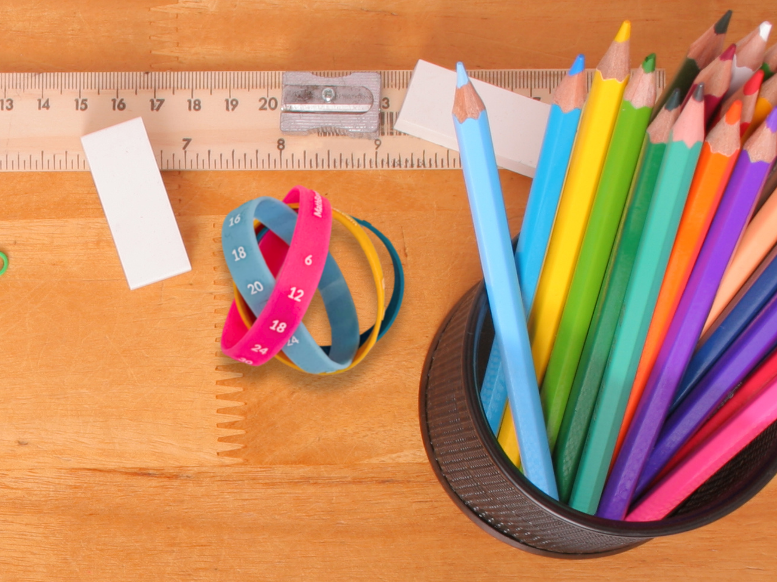 A collection of colorful pencils in a cup, a blue and pink measuring tape, a white sticky note, a wooden ruler, and a small white rectangular object on a wooden surface.