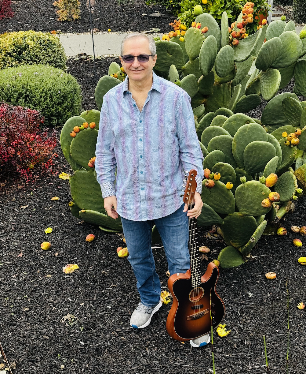 A man wearing sunglasses, a patterned button-up shirt, and jeans standing in front of a large prickly pear cactus with orange fruit, holding an acoustic guitar in a landscaped garden with various bushes and flowers.