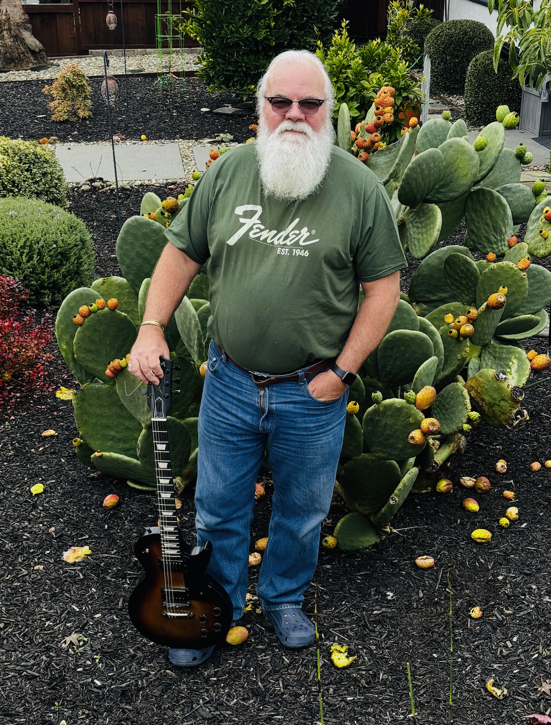 A man with white hair and a beard standing outdoors in a garden with prickly pear cacti and various green plants. He's wearing sunglasses, a green Fender T-shirt, jeans, and Crocs, and holding a small electric guitar.