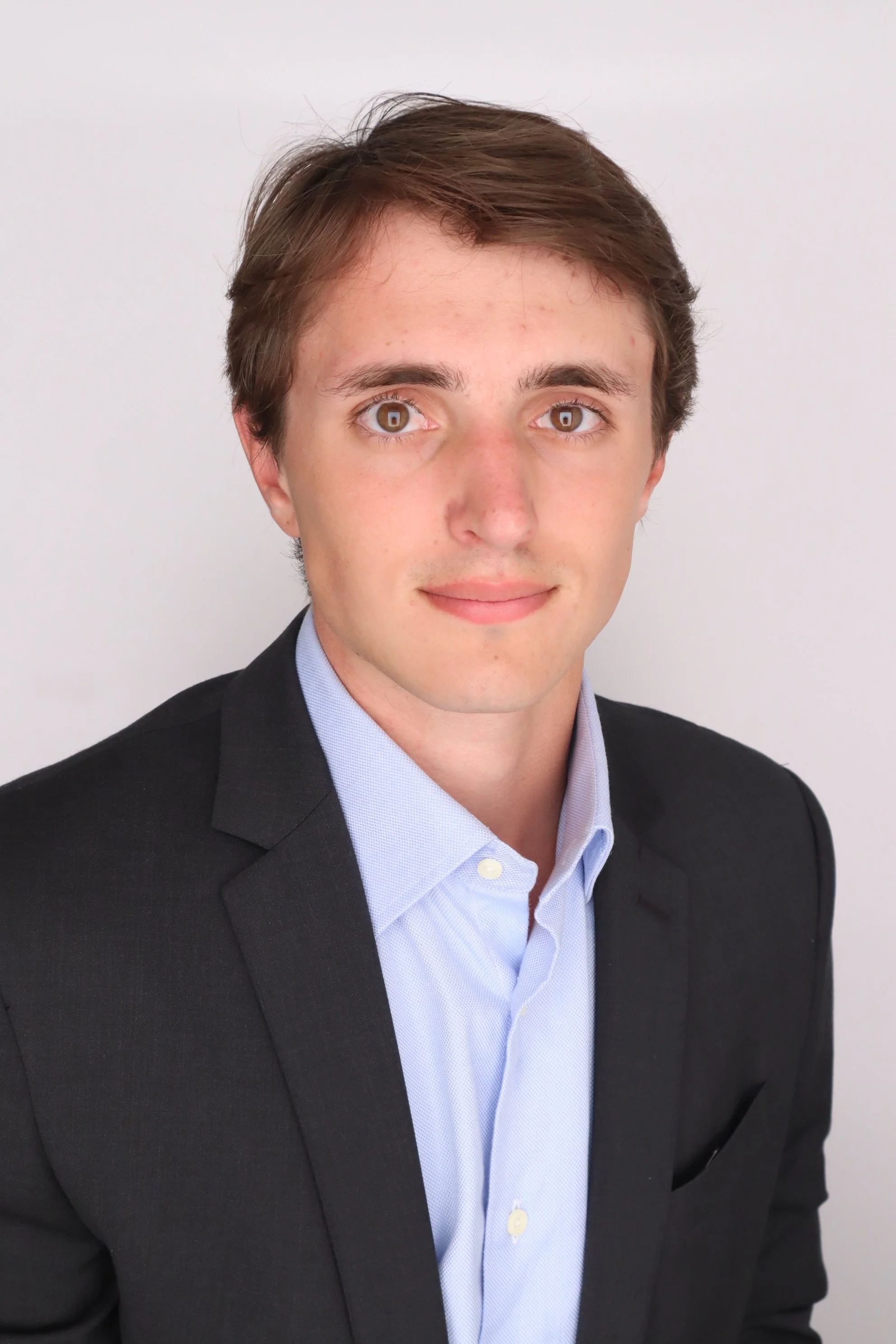 A young man with brown hair and brown eyes, wearing a black suit jacket and light blue dress shirt, posing against a plain white background.