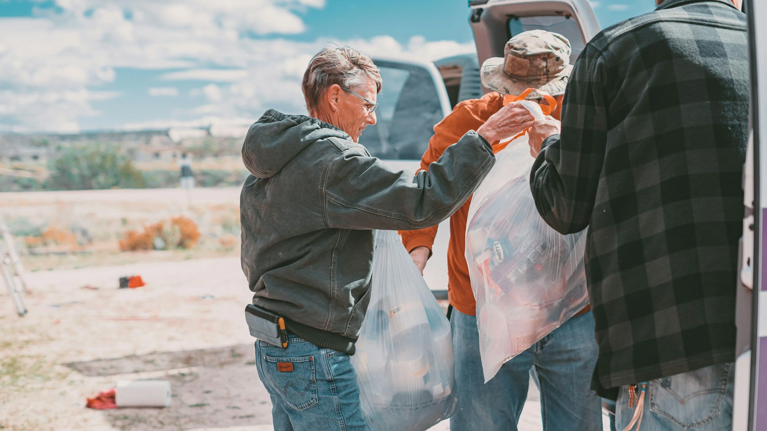 Three people outdoors loading food into plastic bags from a van, with a clear, sunny sky and desert landscape in the background.