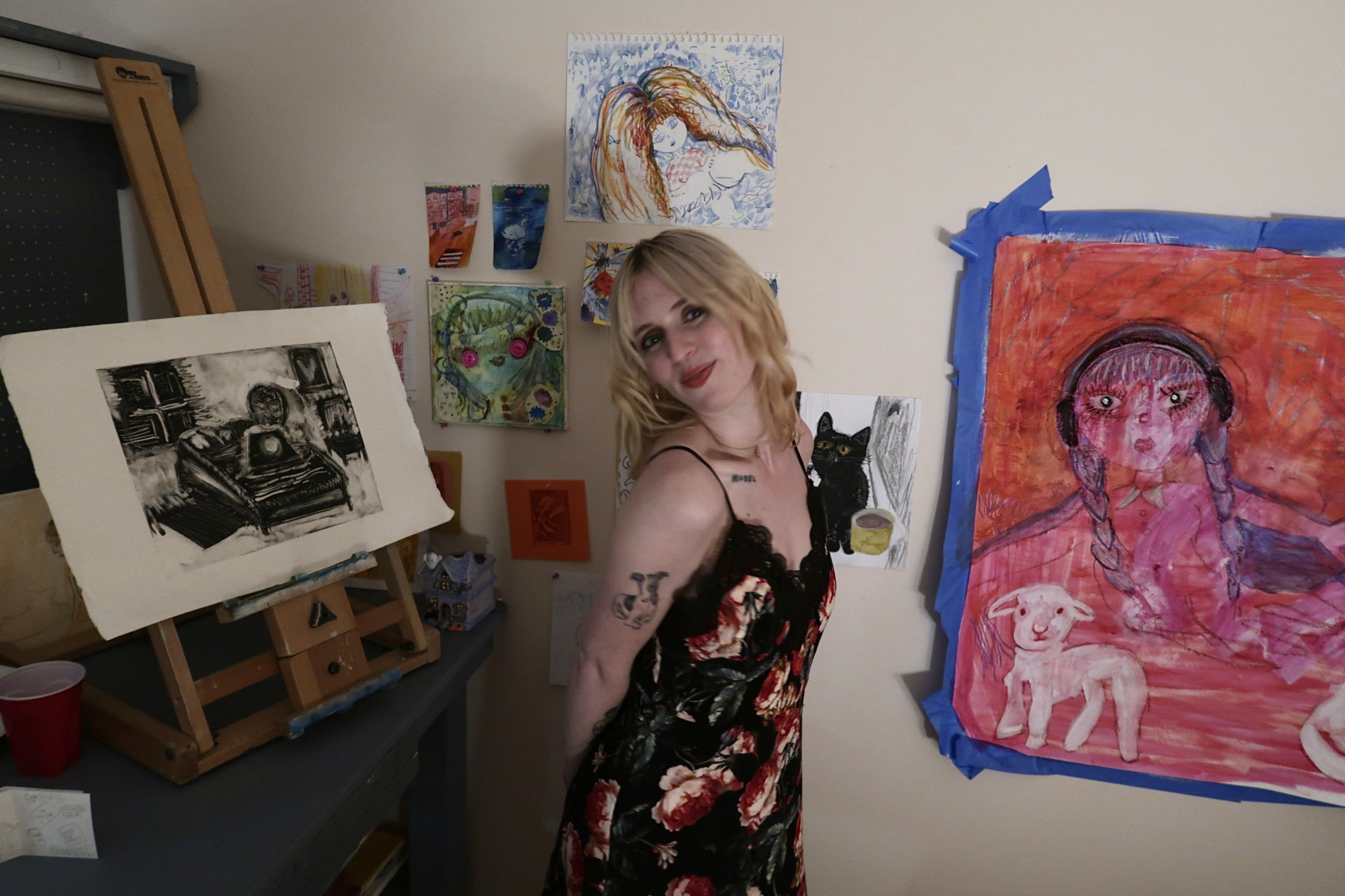 A woman with blonde hair and tattoos standing in front of colorful artwork on a wall, including a large red painting of a girl with braids and headphones, a small black cat drawing, and various smaller paintings and prints in a studio or art space.
