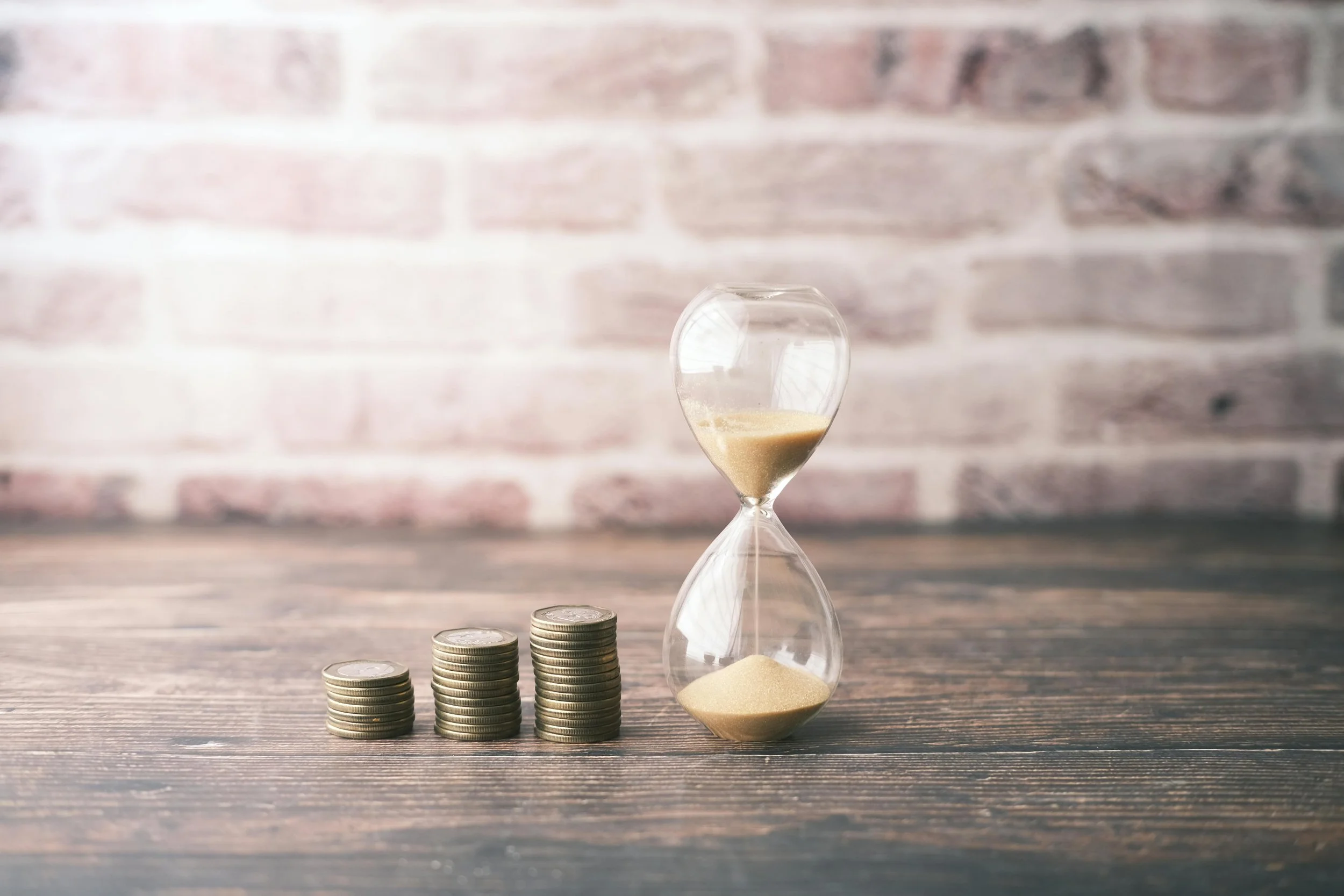 Stacks of coins and an hourglass filled with sand on a wooden surface against a brick wall background.