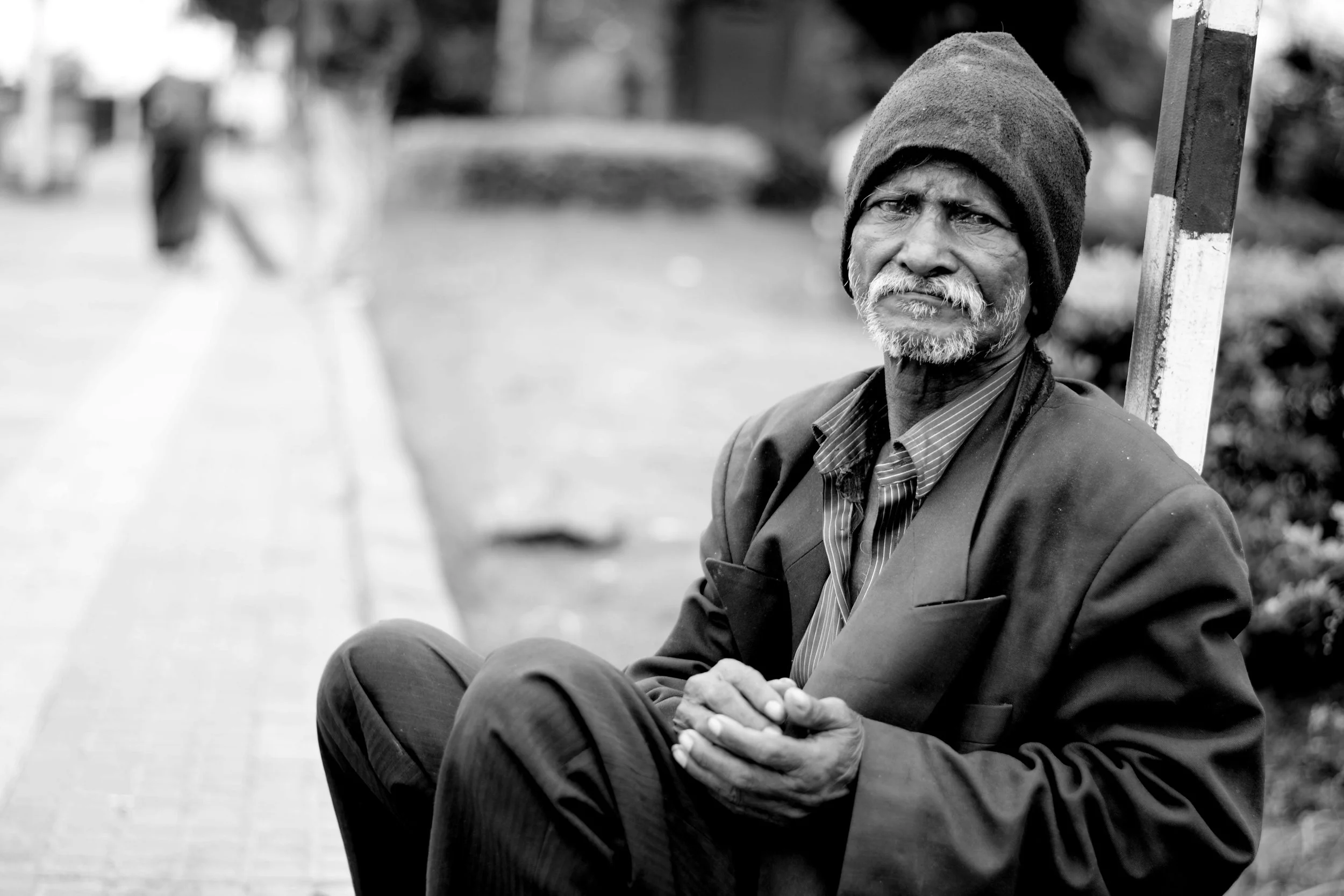 An elderly man with a beard wearing a hat and a suit jacket sitting on a bench outdoors.
