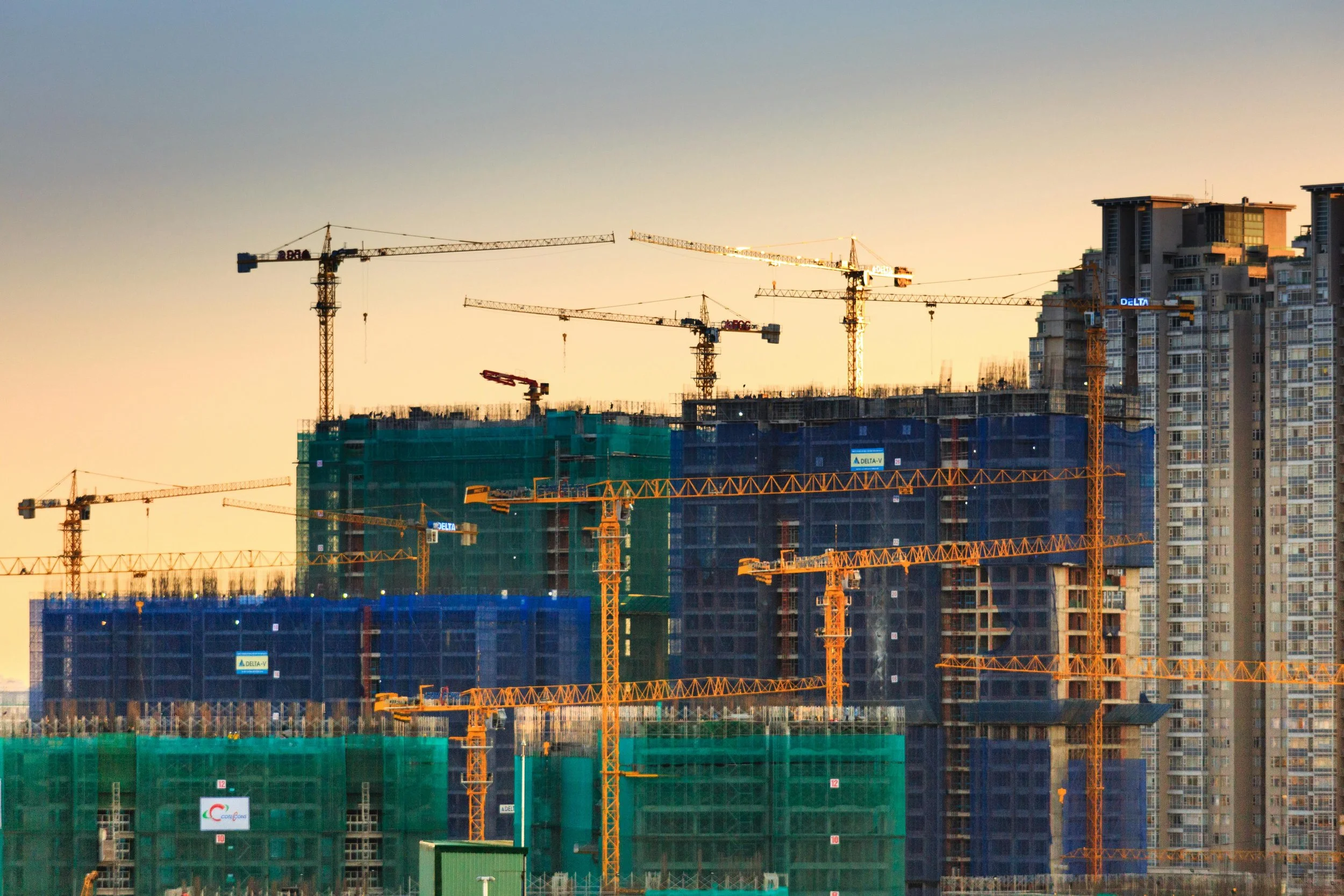 Construction site with multiple orange cranes and partially built high-rise buildings covered in green and blue safety netting during sunset.