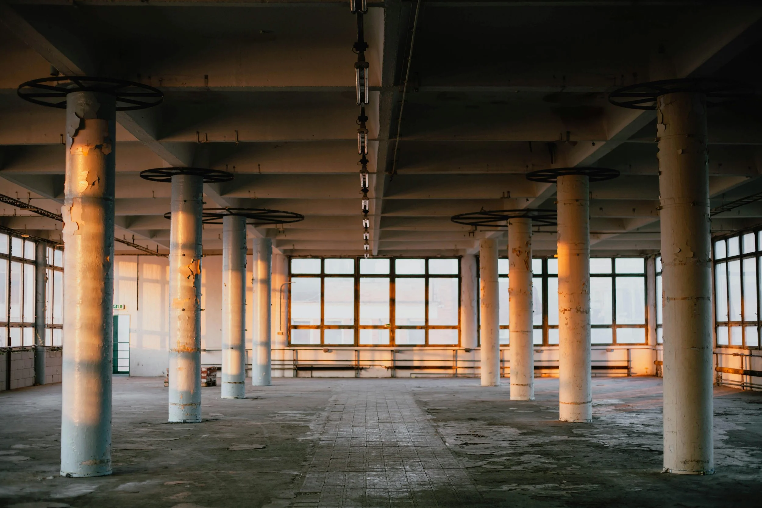 Empty industrial space or warehouse with white columns, large windows, and concrete flooring, illuminated by warm sunlight from the windows.
