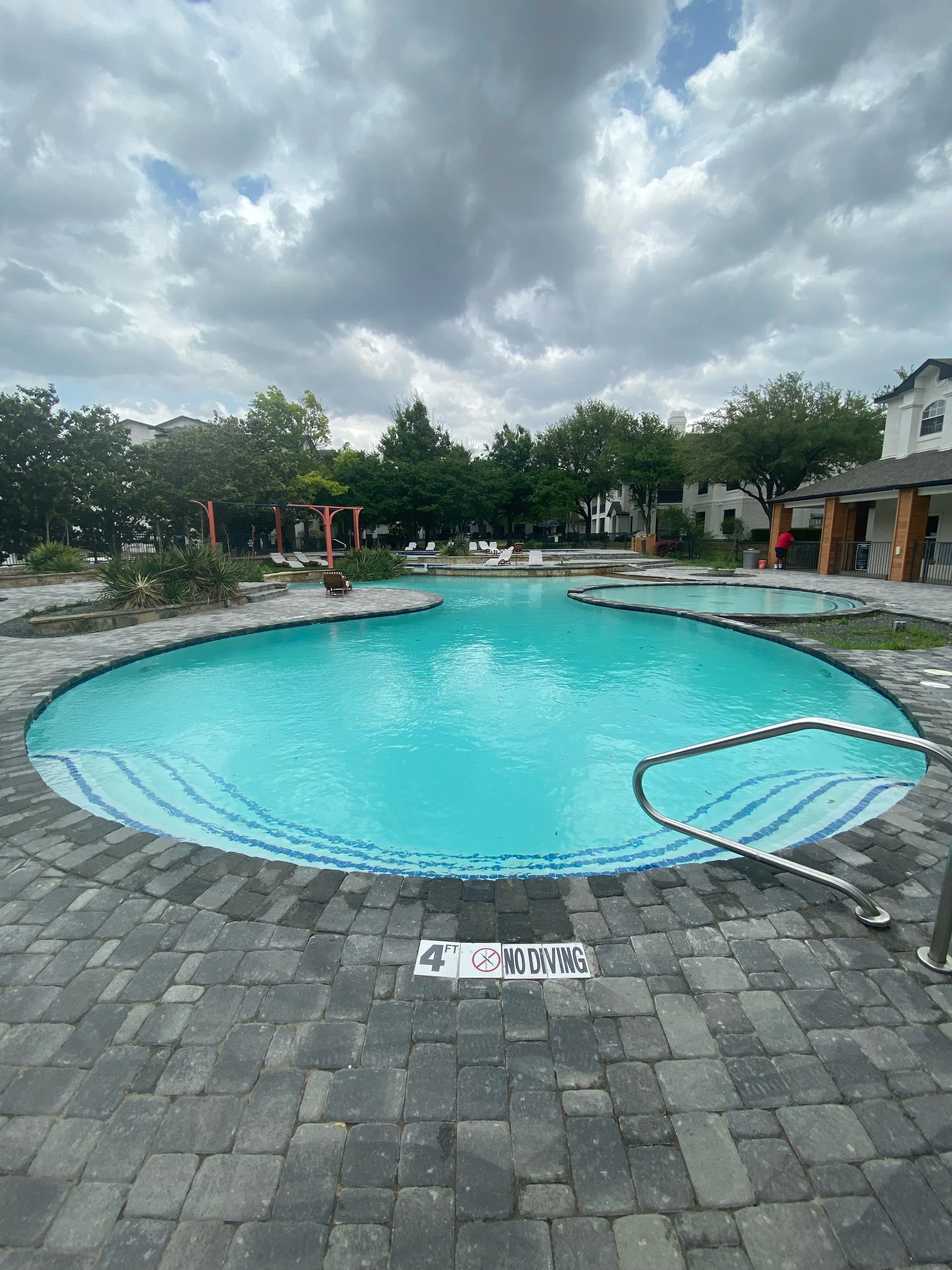 Swimming pool area with a sign on the ground that reads '4 FT. NO DIVING', pool ladder, surrounding patio with chairs, trees, and a cloudy sky.