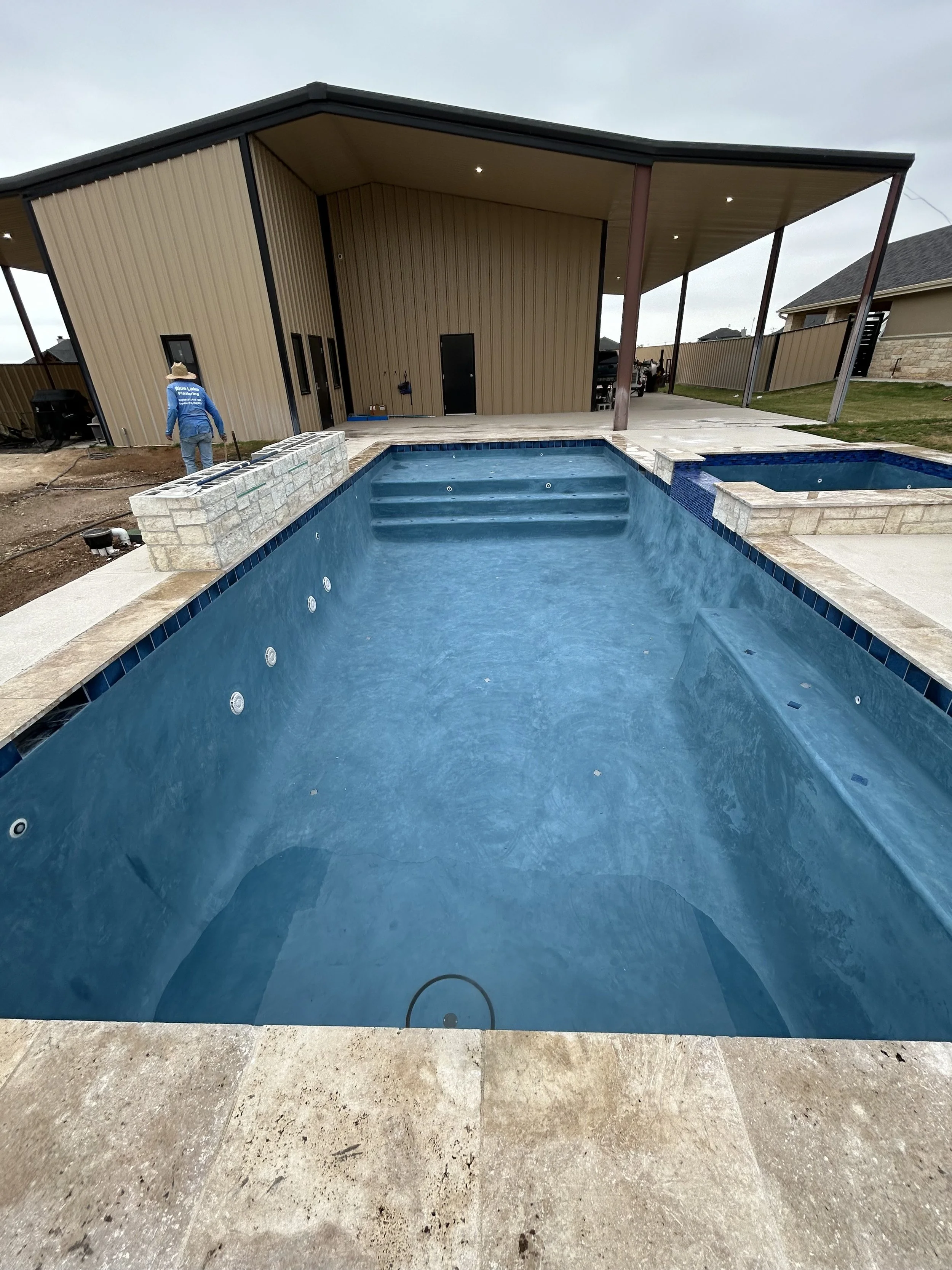 A backyard with an in-ground swimming pool and a hot tub, with a covered patio area in the background, during daytime with cloudy skies.