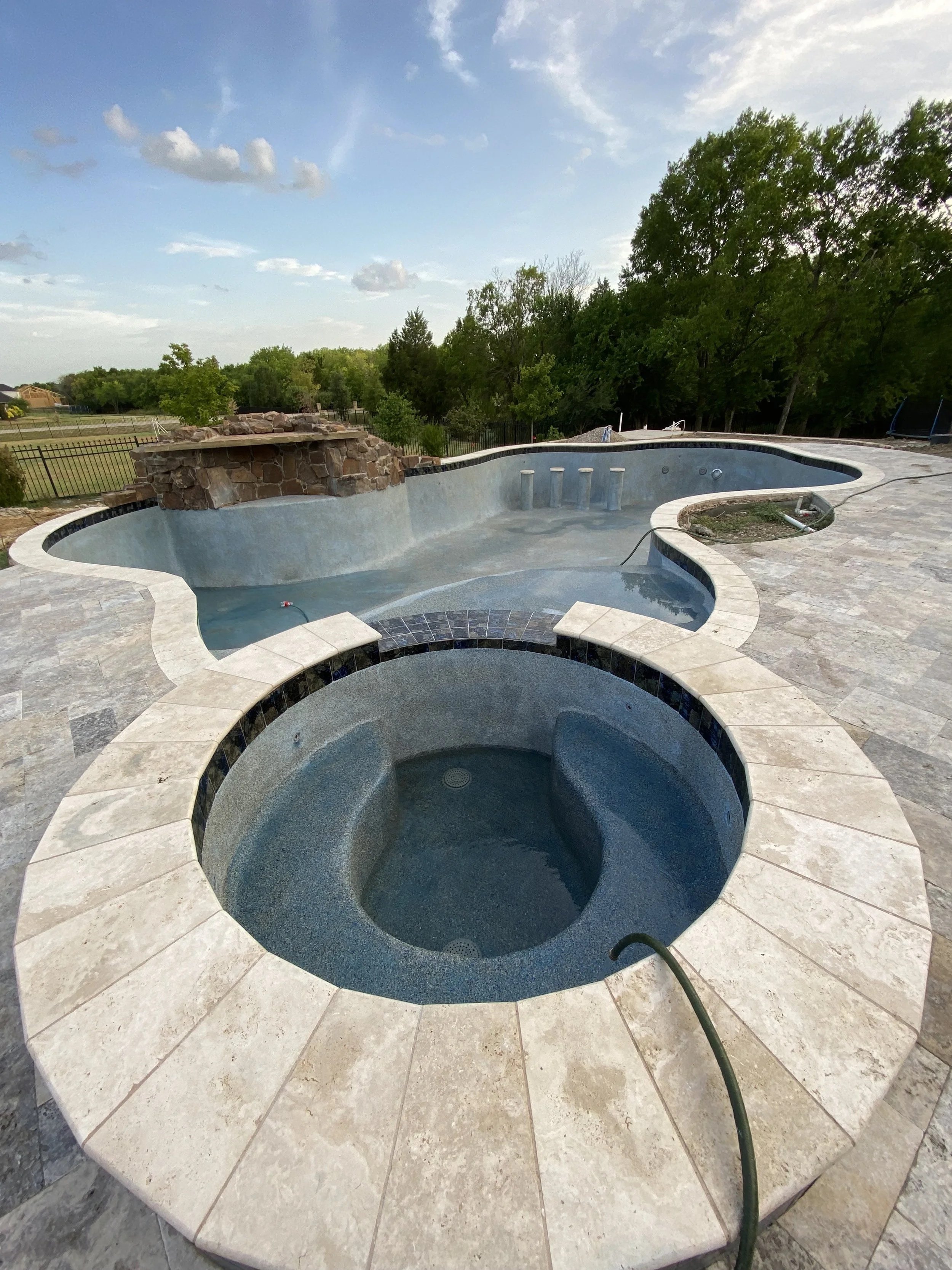 Image of an unfinished pool area with two circular hot tubs and a larger swimming pool, surrounded by stone paving, with a natural landscape of trees and a partly cloudy sky in the background.