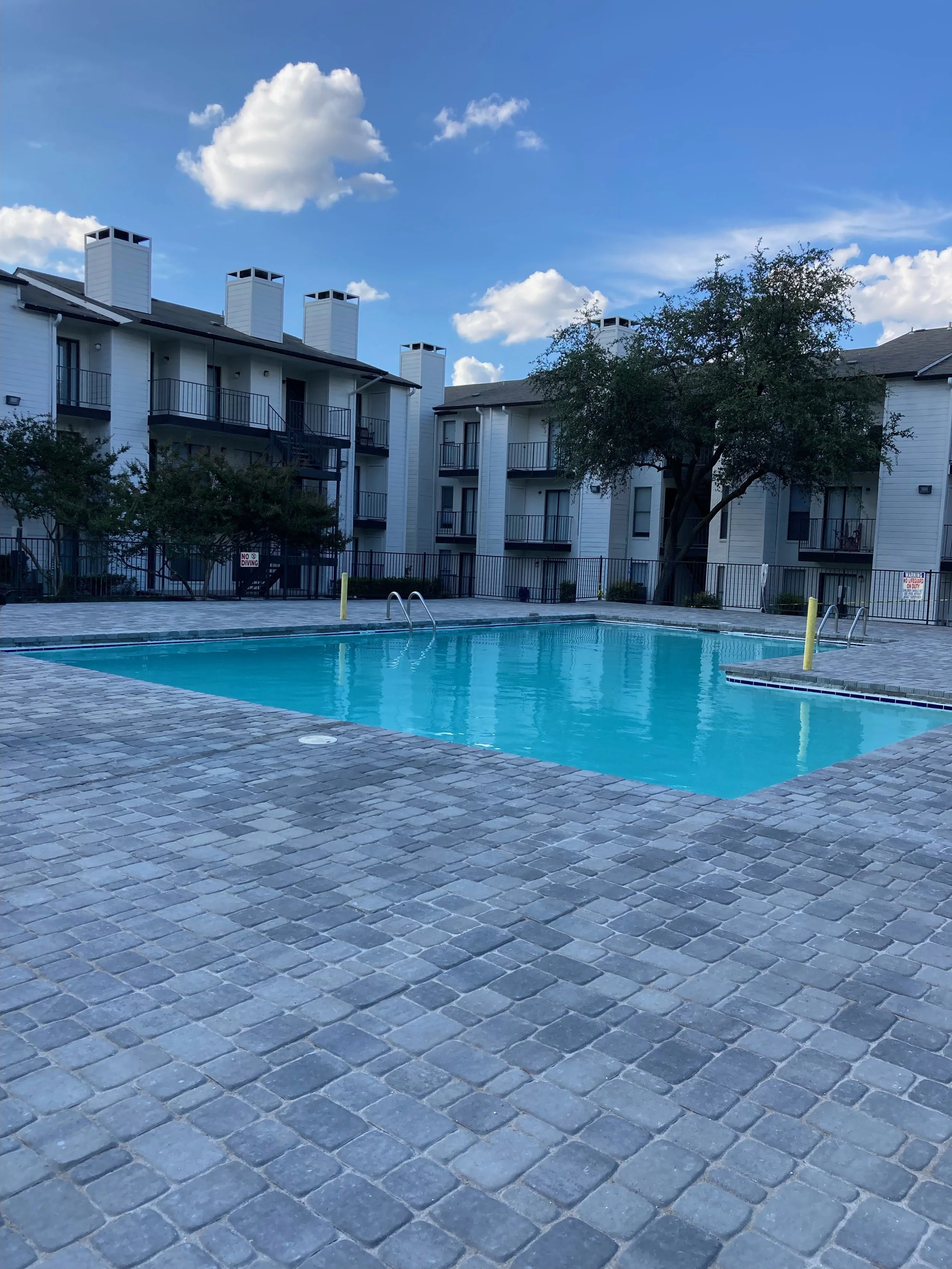 Apartment complex with a swimming pool in the foreground, surrounded by a brick patio. The building has multiple floors, white siding, and several chimneys on the roof. The sky is blue with scattered clouds.