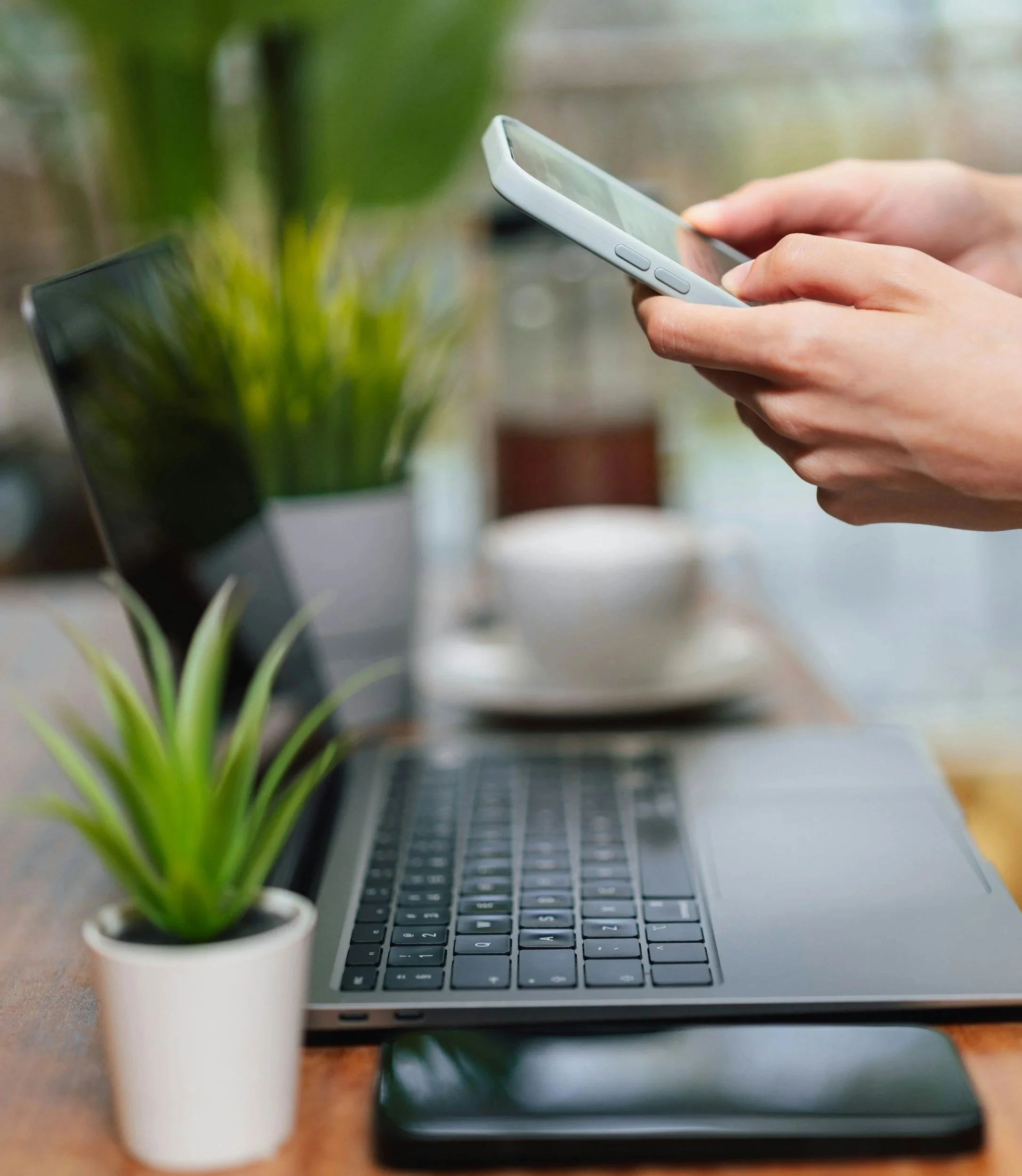 Close-up of hands holding a smartphone with a silver laptop, small potted plant, and black smartphone on a wooden table, with a blurred background.