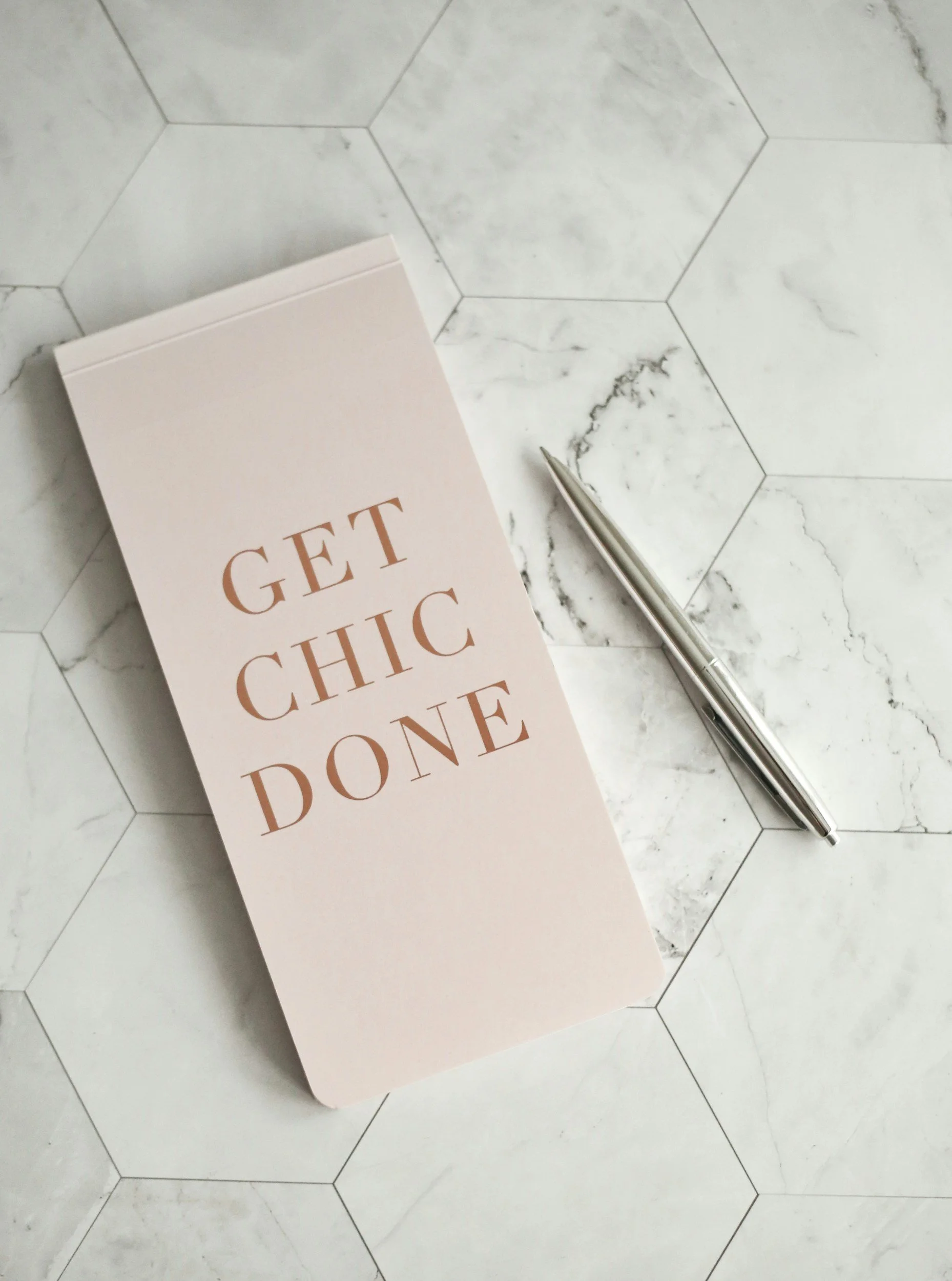 A light pink notebook with the phrase "Get Chic Done" in uppercase letters on the cover, lying on a white marble countertop with gray veining and hexagonal tiles. A silver pen is placed next to the notebook.