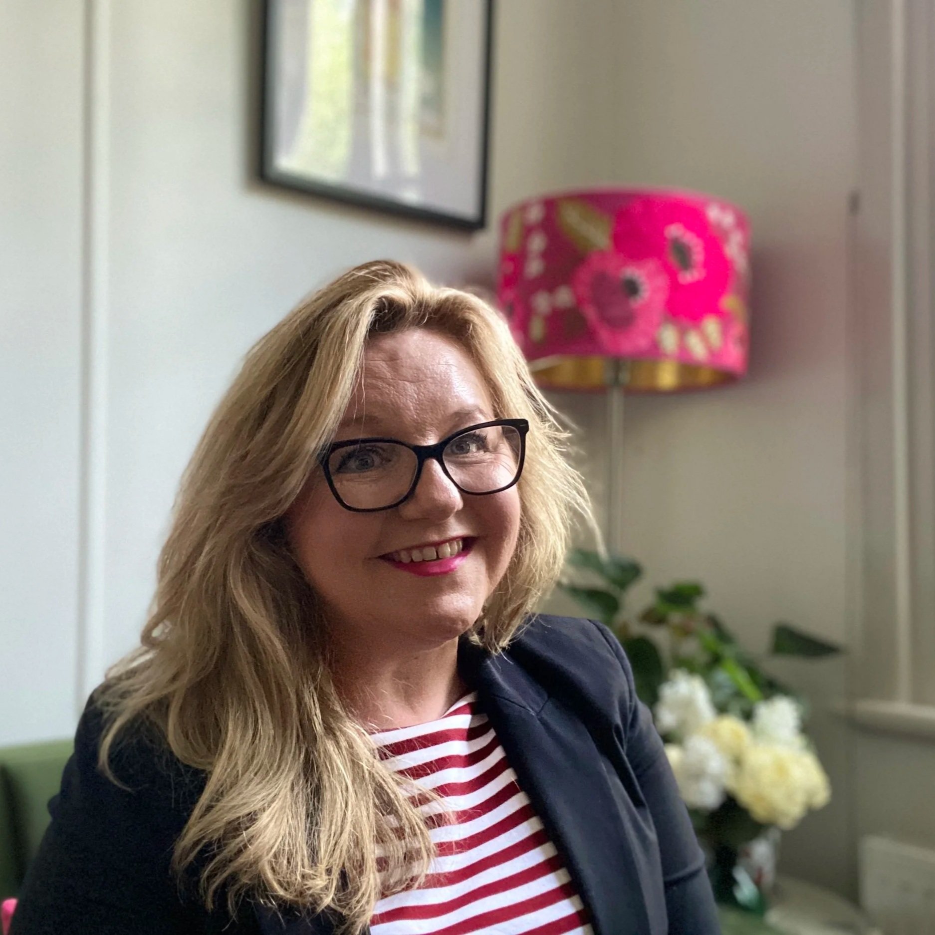A woman with long blonde hair, glasses, and red lipstick smiling at the camera, seated in a room with a pink floral lamp and white flowers in the background.