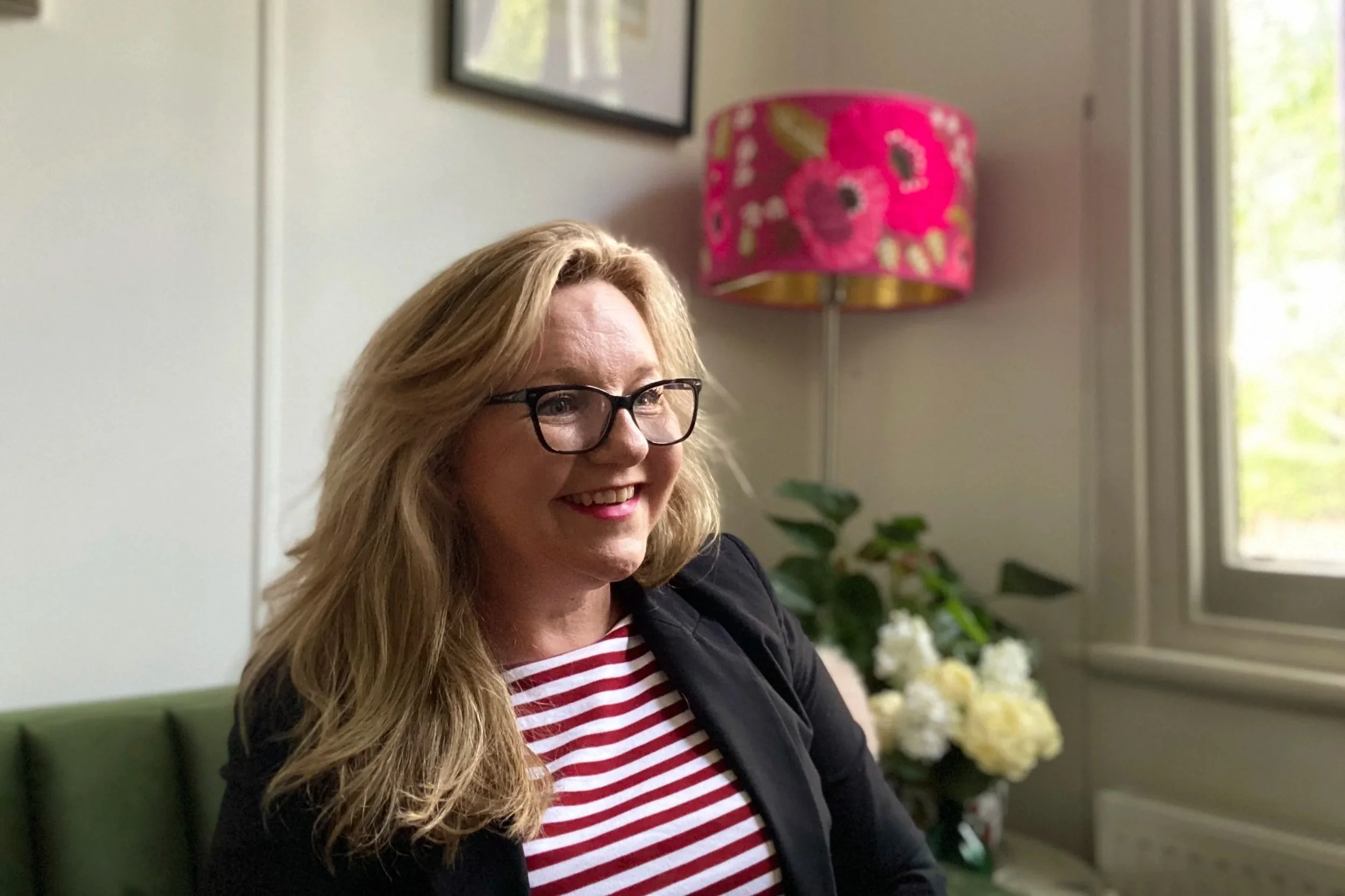 A woman with long blonde hair, glasses, and red lipstick, smiling at the camera. She is wearing a black blazer over a red and white striped shirt, seated in a room with a window, a pink floral lampshade, and a vase of white flowers in the background.