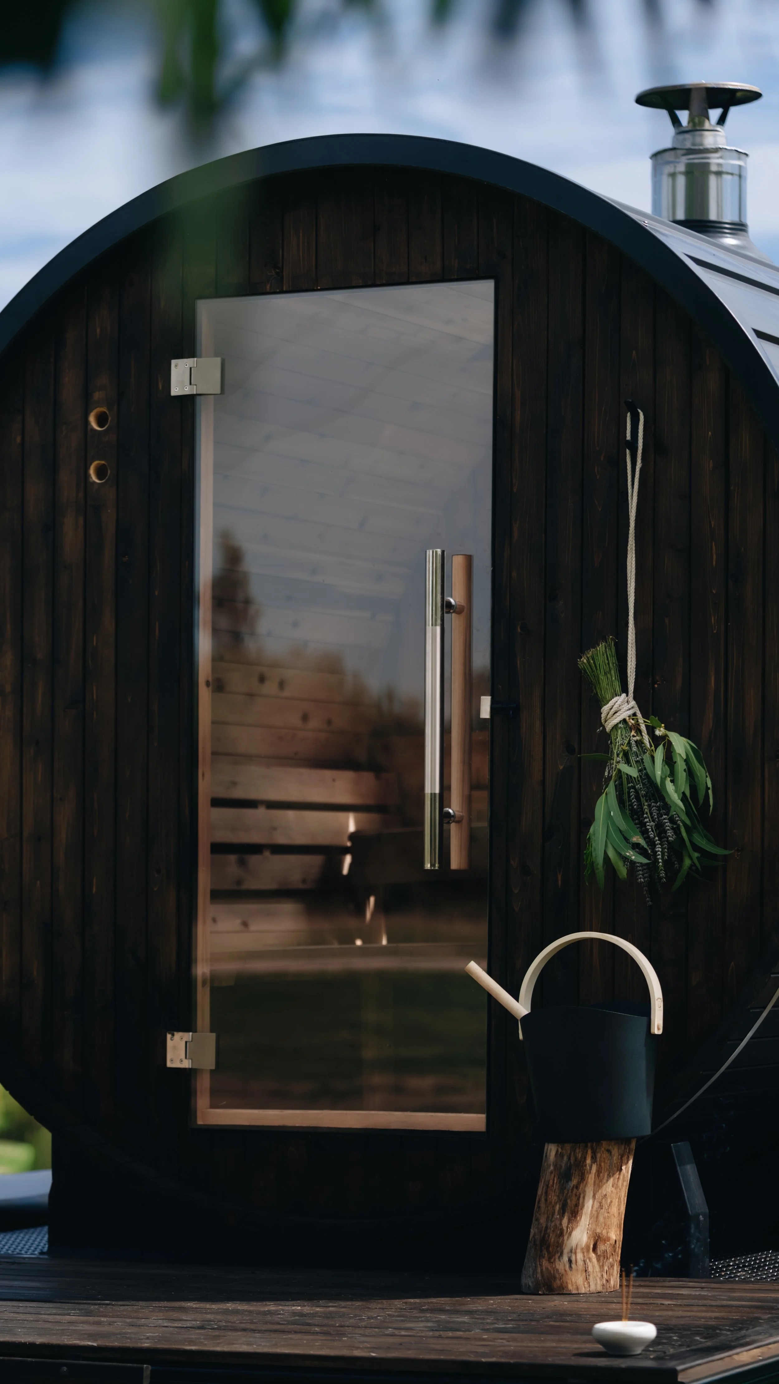 A small black sauna with a glass door, a wooden interior, and a metal chimney on top. Outside, there is a wooden deck with a black watering can and a white ceramic bowl with a stick and a bundle of greenery tied with string hanging outside the sauna.