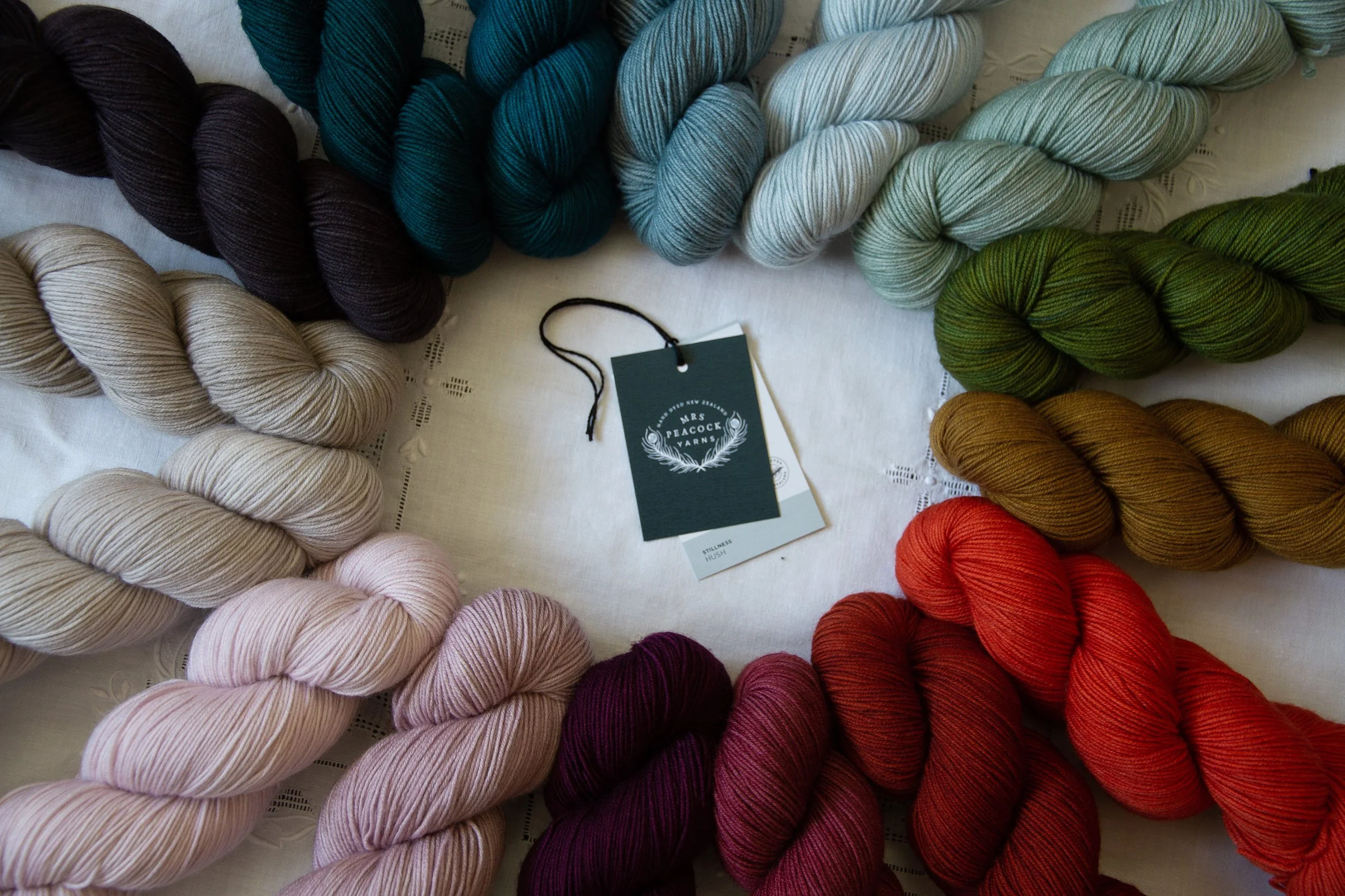 Multiple skeins of colorful yarn arranged in a circle on a white table, with a Mrs. Peacock Yarns tag in the center.