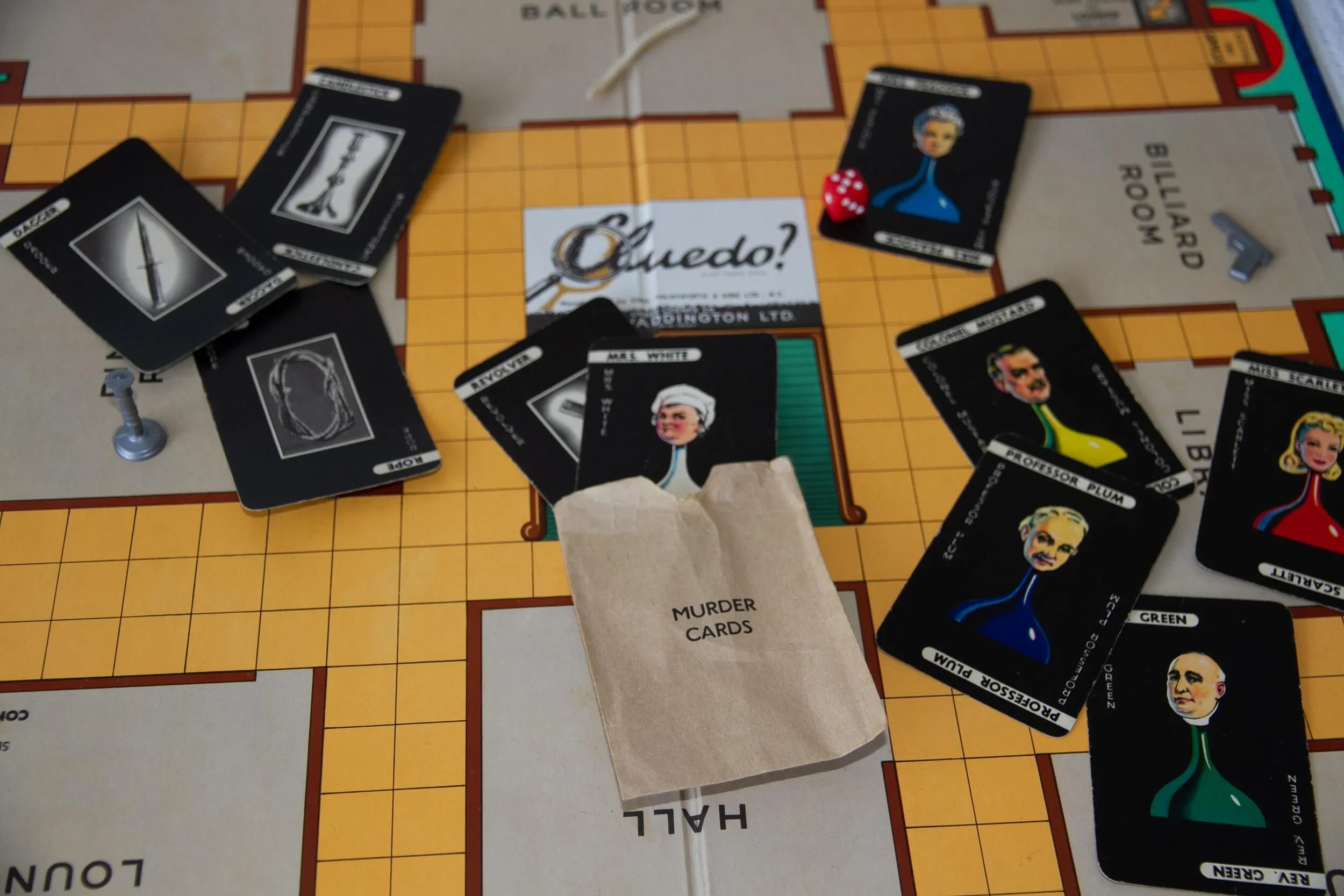 Cluedo board game with character cards, a small paper bag labeled 'Murder Cards', and game pieces on a yellow grid game board.