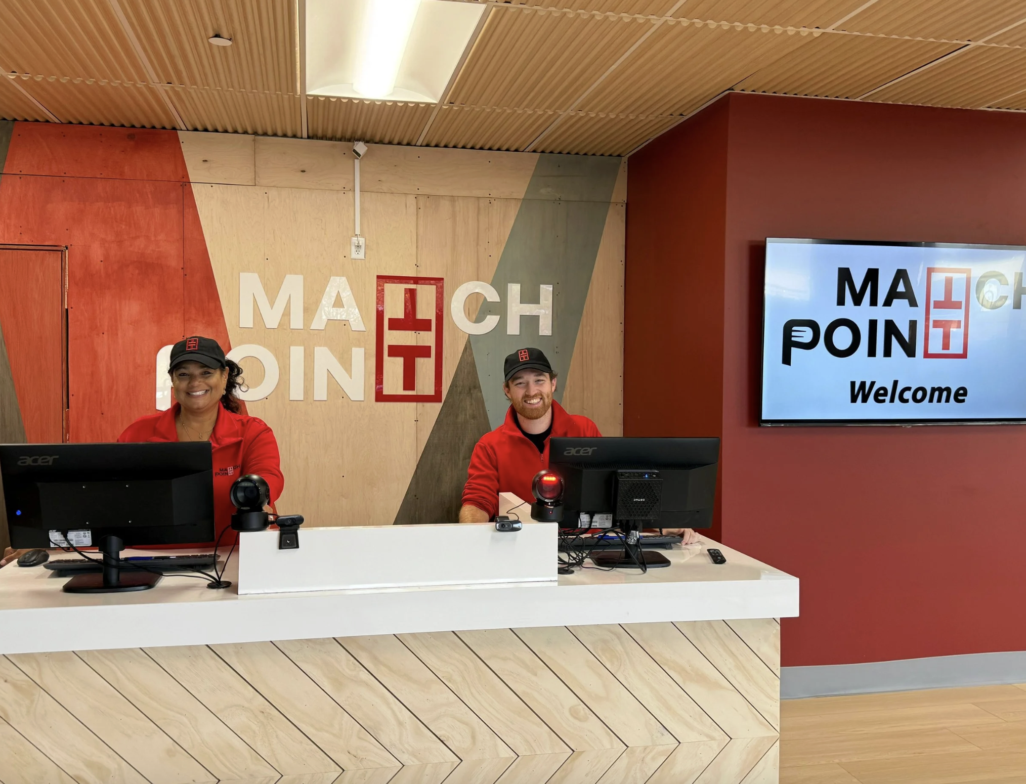 Two staff members, a woman and a man, sitting behind a reception desk with computers at a place called Mach Point. They are smiling and wearing red jackets and black caps. Behind them is a wall with the Mach Point logo and a large screen displaying t