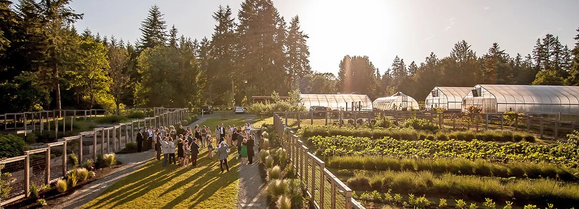 A group of people gathered outdoors on a sunny day in a garden with greenhouses and lush plants, surrounded by tall trees.