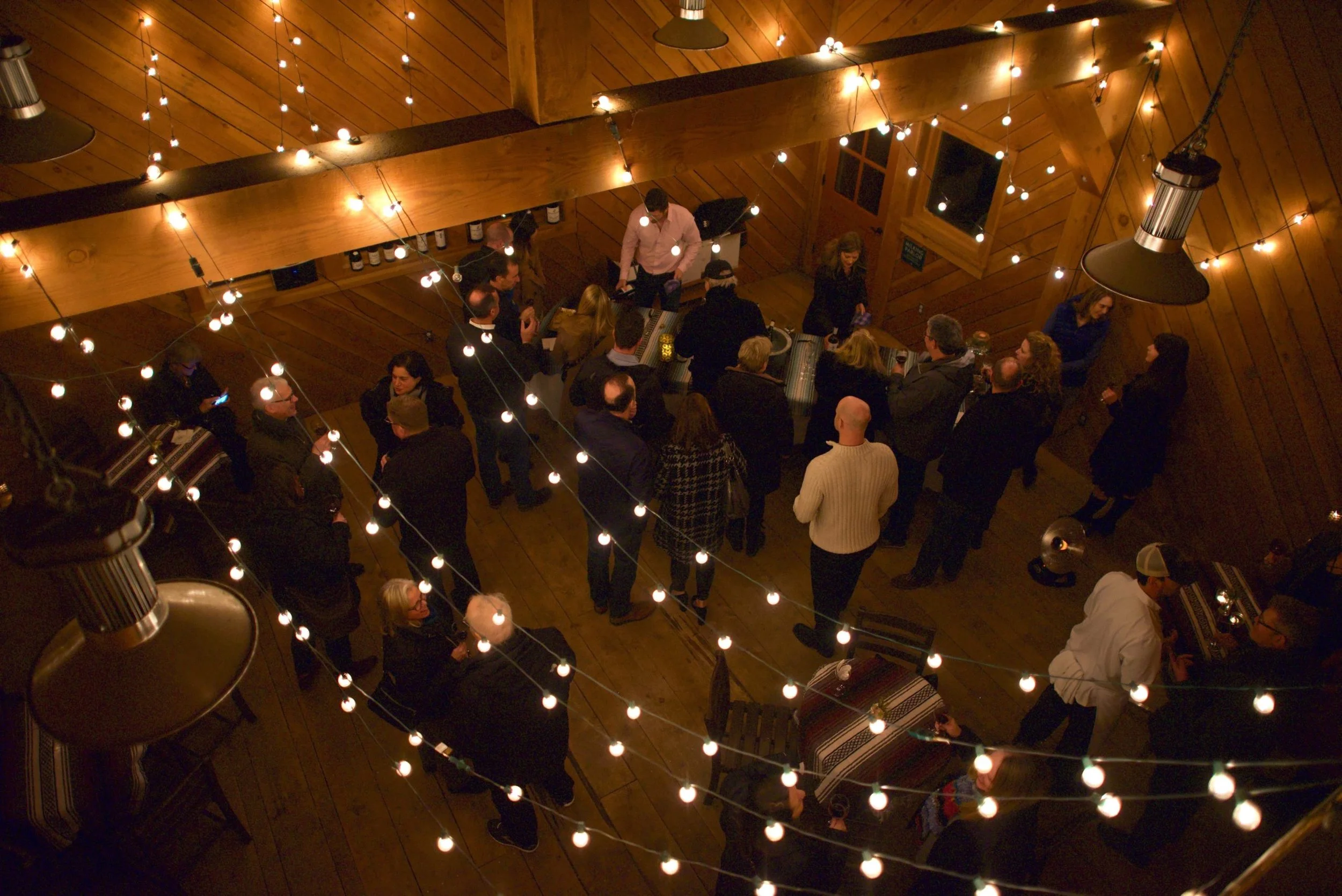 People mingling at a social gathering in a warmly lit barn with string lights and table setups.