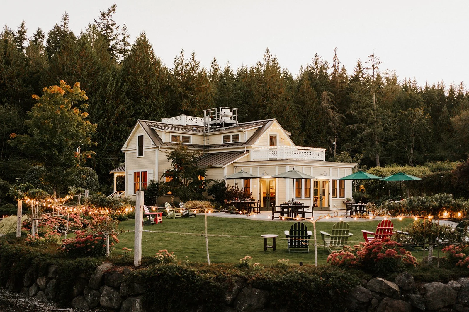 Large white house with multiple stories and a metal roof, surrounded by a well-kept lawn and garden, with string lights, outdoor tables with umbrellas, and colorful Adirondack chairs, backed by a forested hillside.
