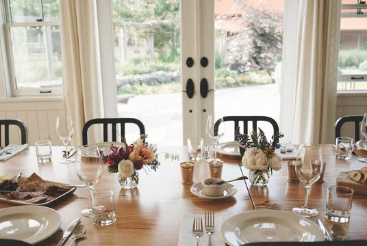 A dining table set with plates, glasses, utensils, and floral centerpieces in a bright room with large windows
