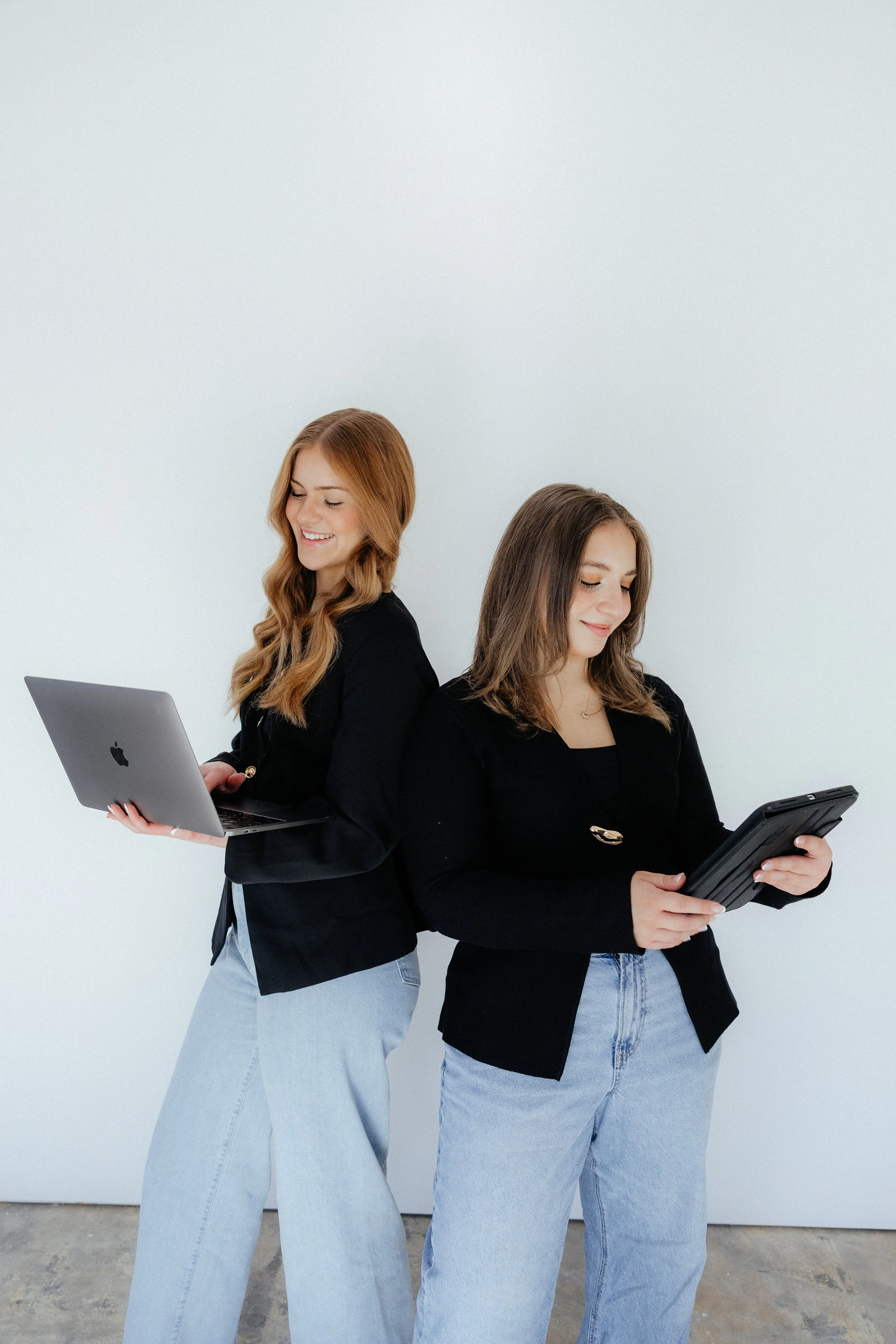 Two women standing back to back, one with a laptop and the other with a tablet, smiling and looking down at their devices.