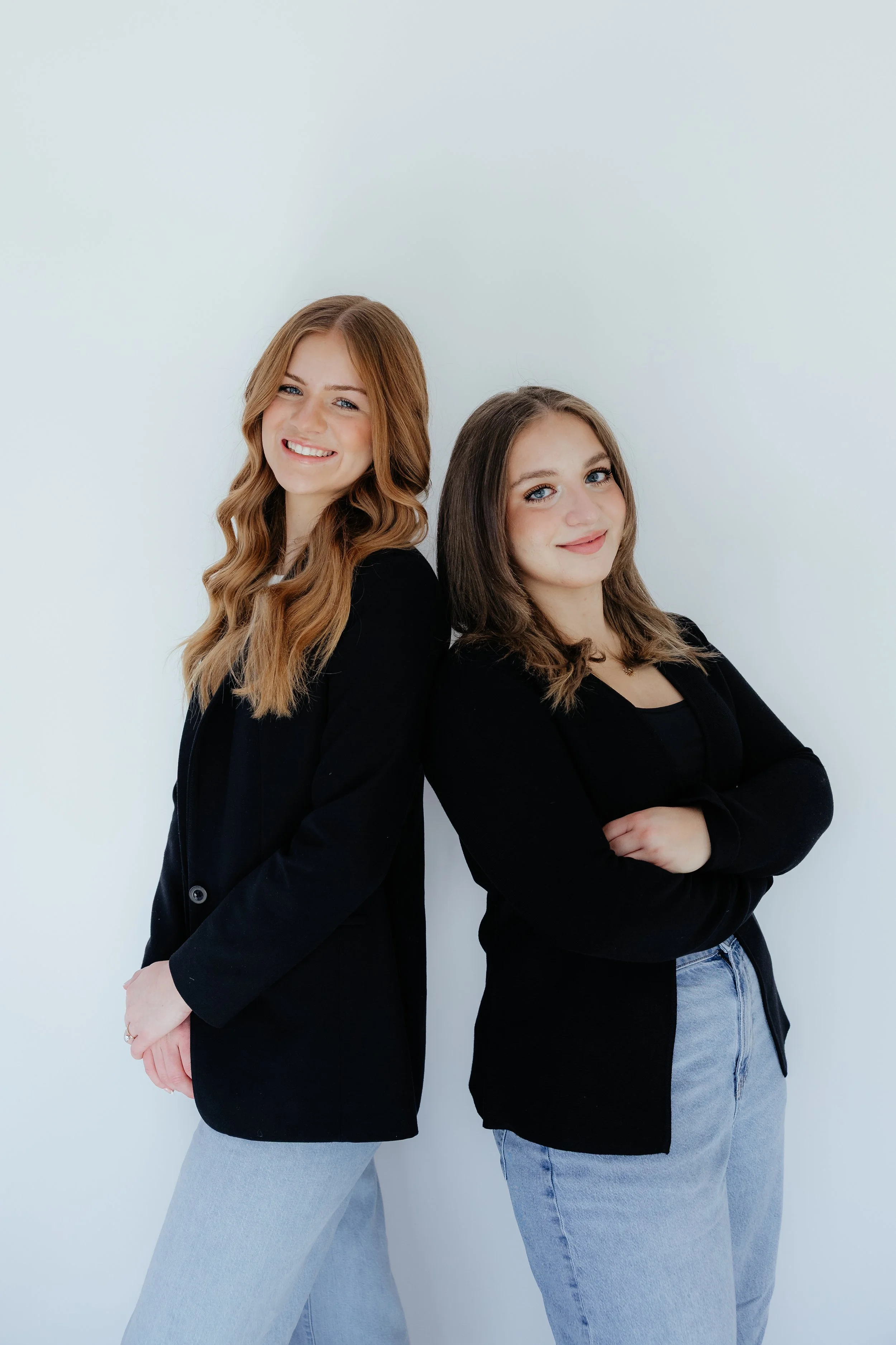 Two young women standing back-to-back, smiling, against a plain light background.